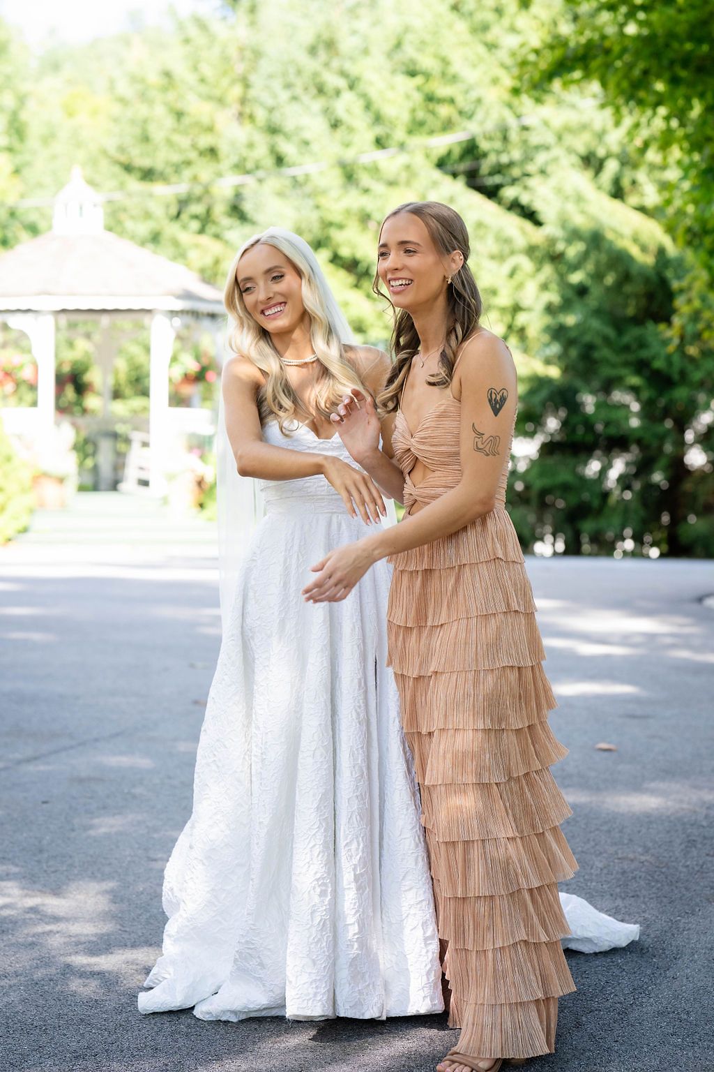 Laughing bride in white gown embraces bridesmaid in tiered champagne dress in sunny garden setting.