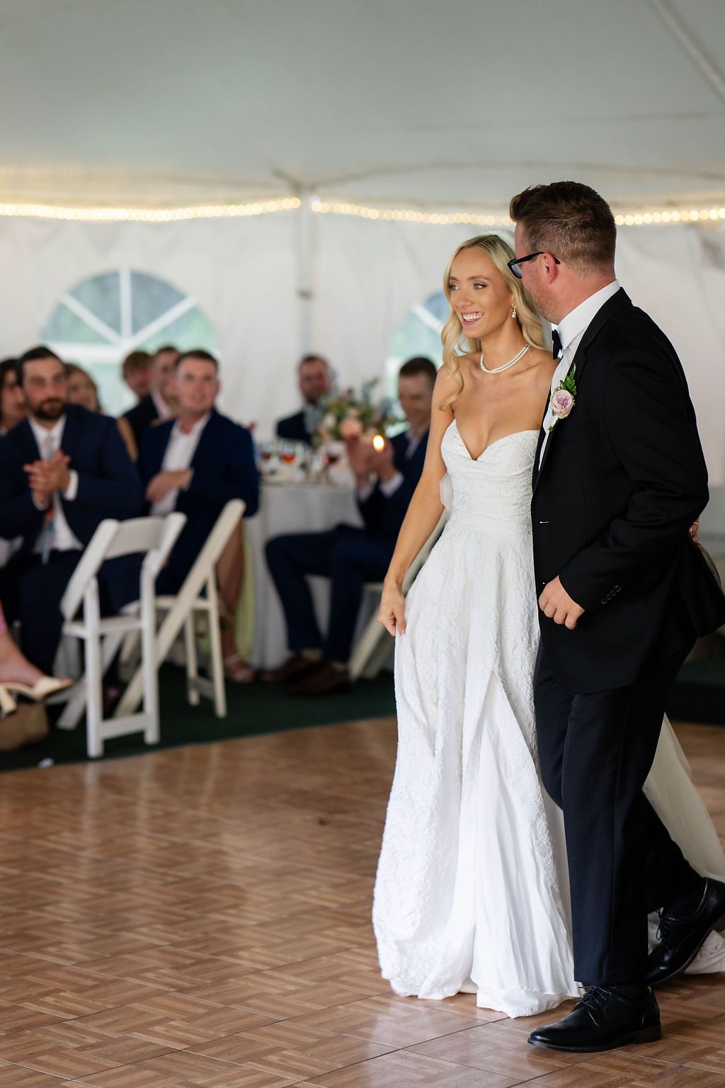 Smiling bride and groom share first dance on wooden floor at tented wedding reception.