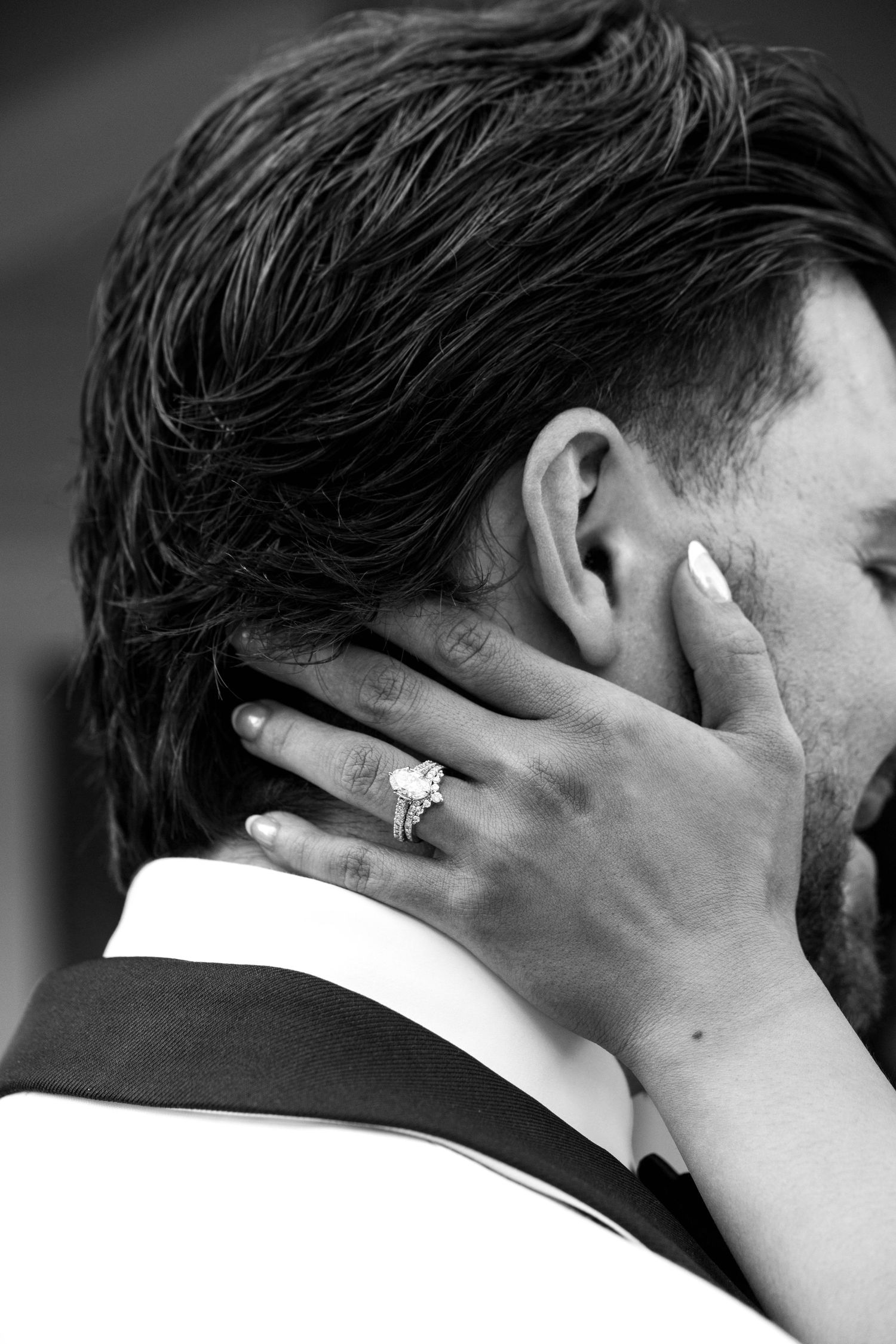 Black and white close-up of bride's hand with diamond ring gently cradling groom's neck during embrace.