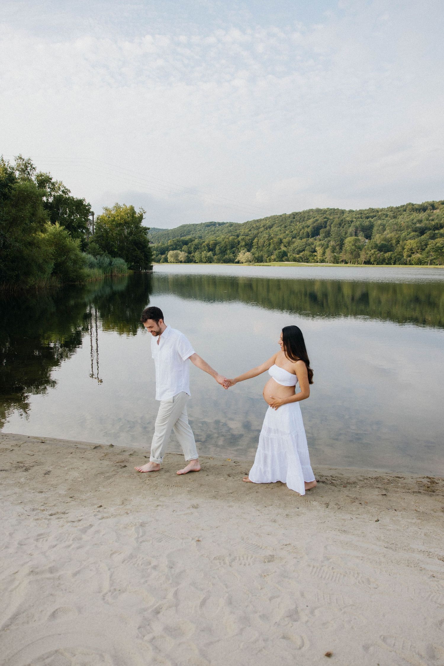 Couple in white outfits holding hands on sandy beach by calm lake with lush green hills in background.