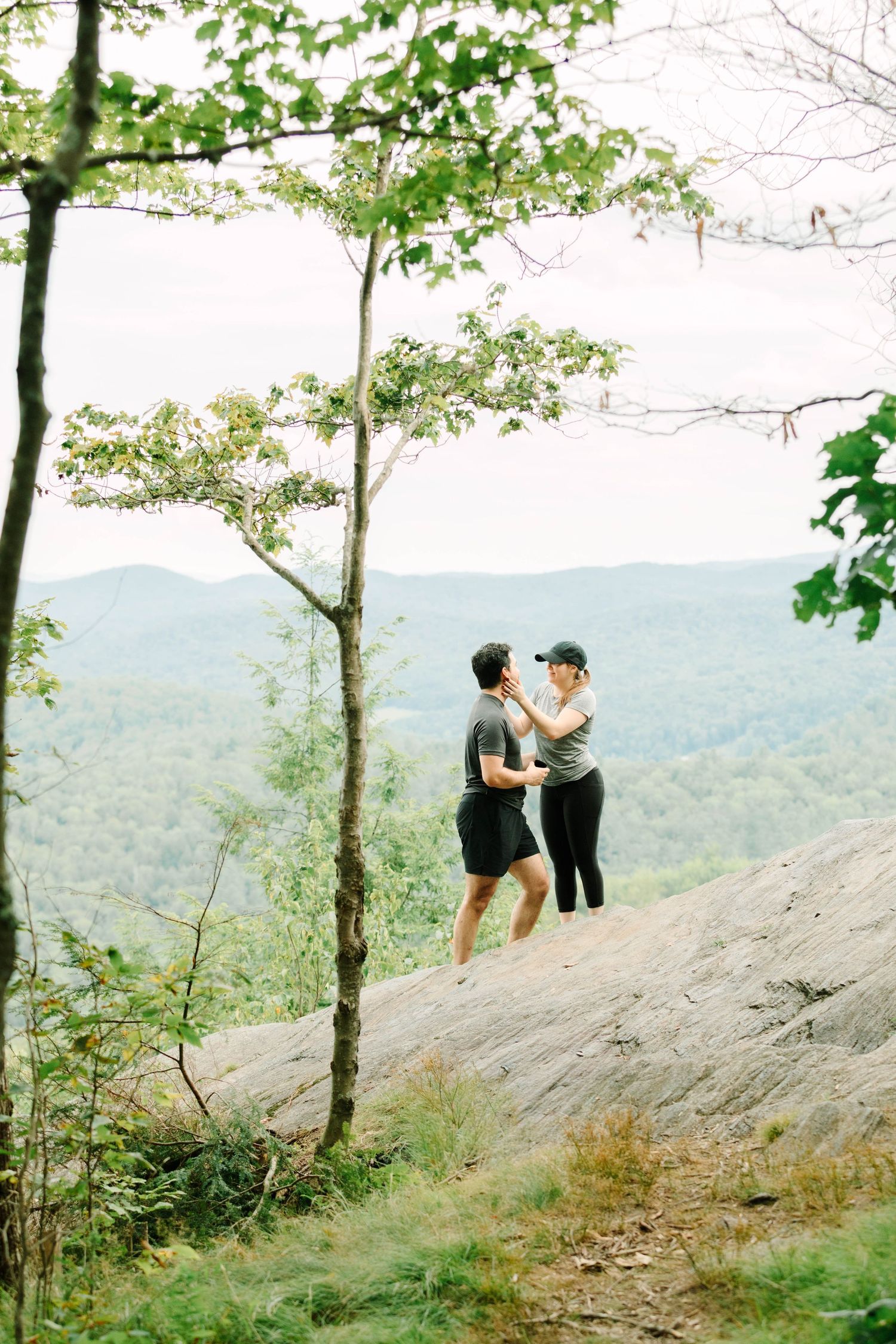 Couple standing on a rocky ledge holding hands with a breathtaking green mountain landscape behind.