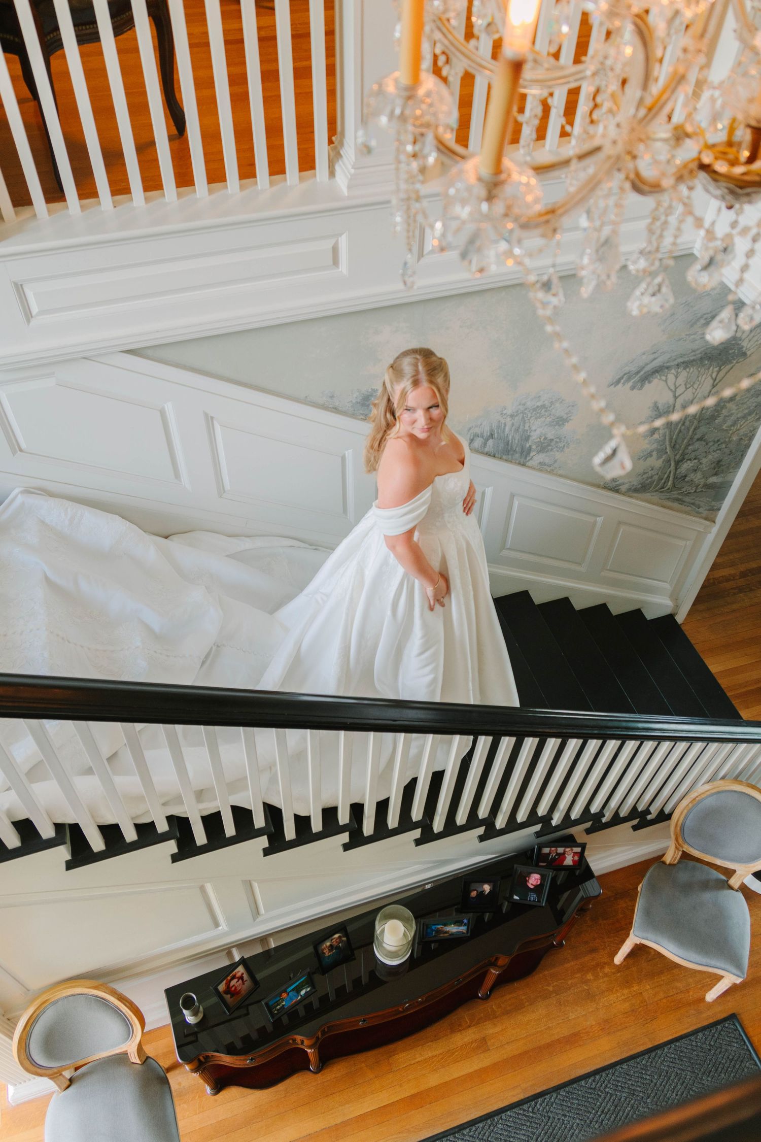 Bride in a white ball gown descends an elegant staircase beneath a crystal chandelier in a grand estate.