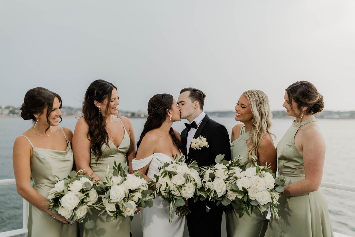 Bride and groom kiss on yacht deck surrounded by four bridesmaids in sage green gowns with bouquets.