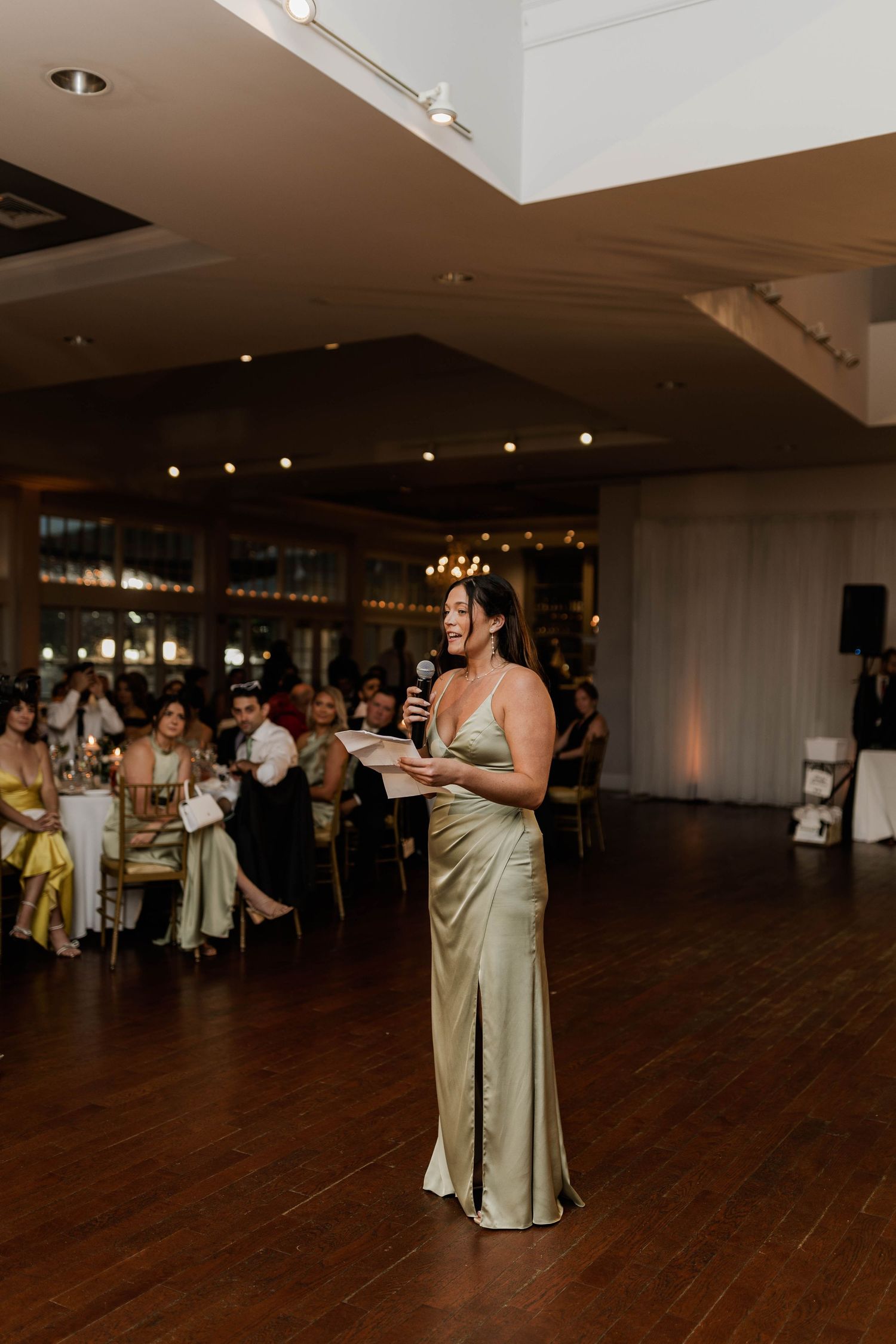 Bridesmaid in sage green satin gown delivers a heartfelt speech at a wedding reception.
