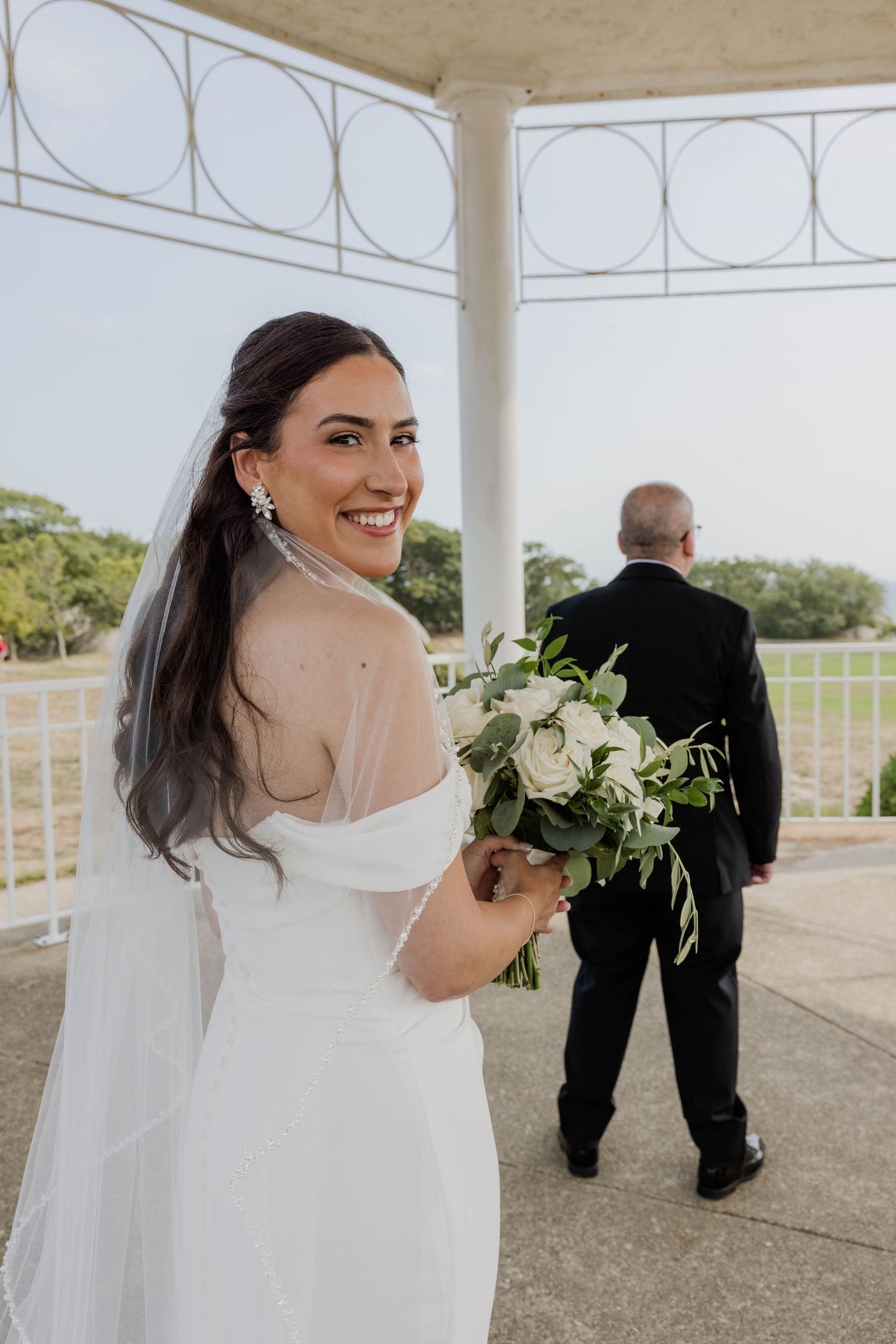 Bride in white off-shoulder gown holding white floral bouquet smiling under gazebo on wedding day.