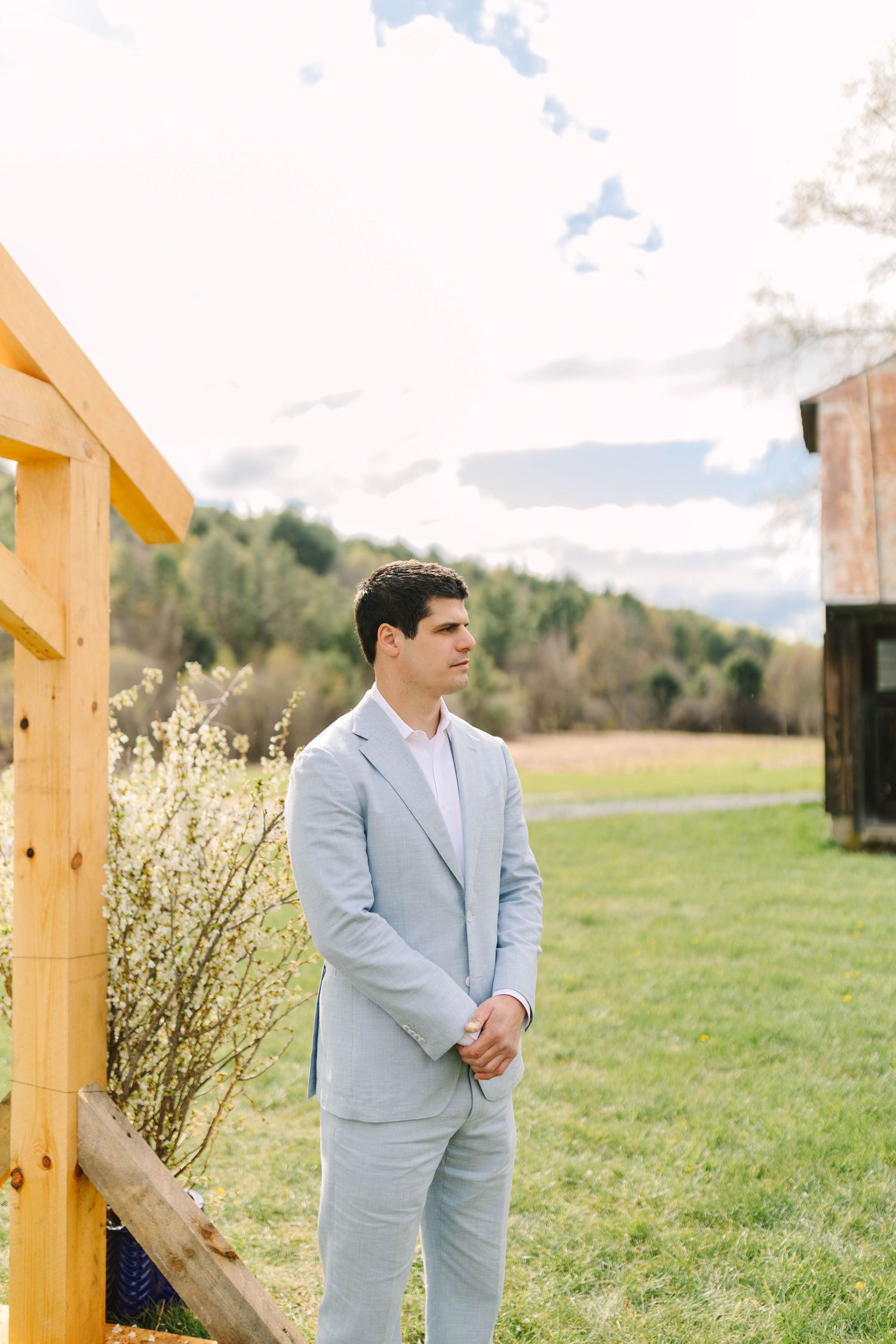 Groom in light blue suit standing outdoors near a wooden fence with barn in background.