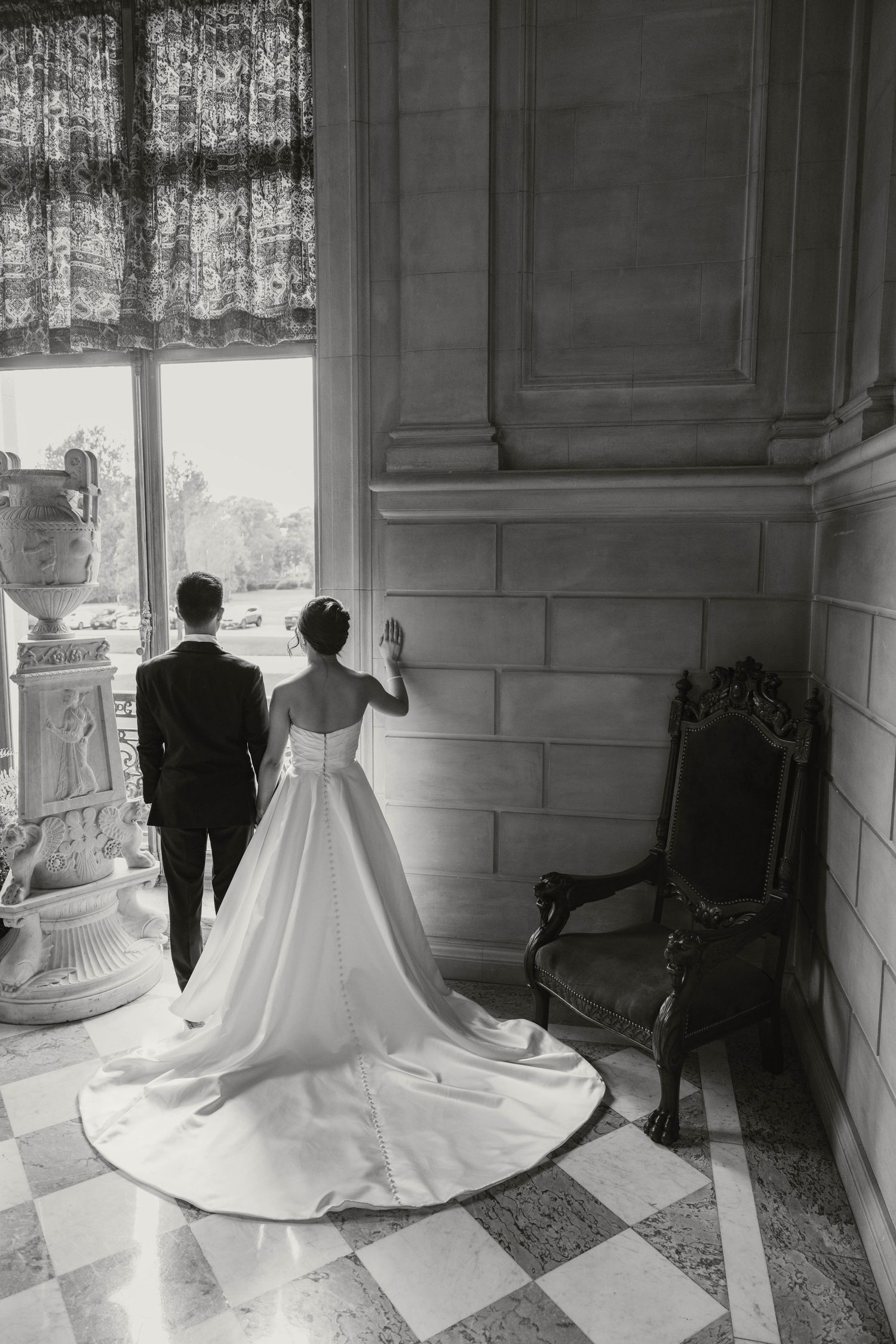 Black and white rear view of bride and groom standing together by tall window in marble-floored hall.