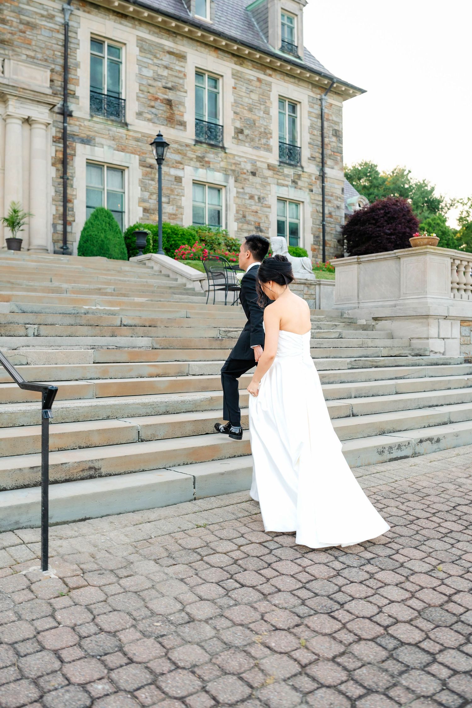 Bride and groom ascend stone steps of elegant manor estate on wedding day.