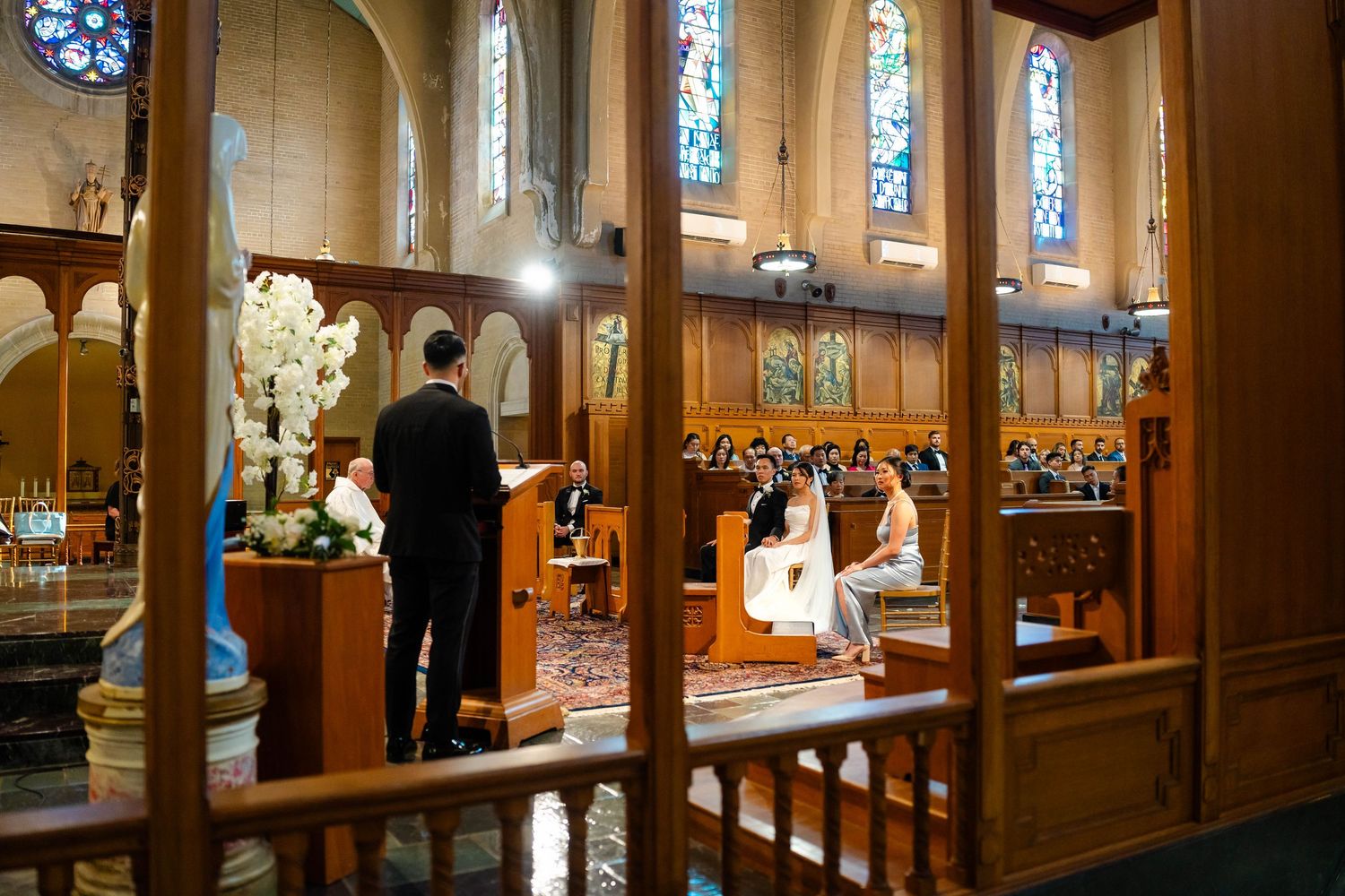 Wedding ceremony viewed through doorway in ornate church with stained glass windows and floral arrangements.