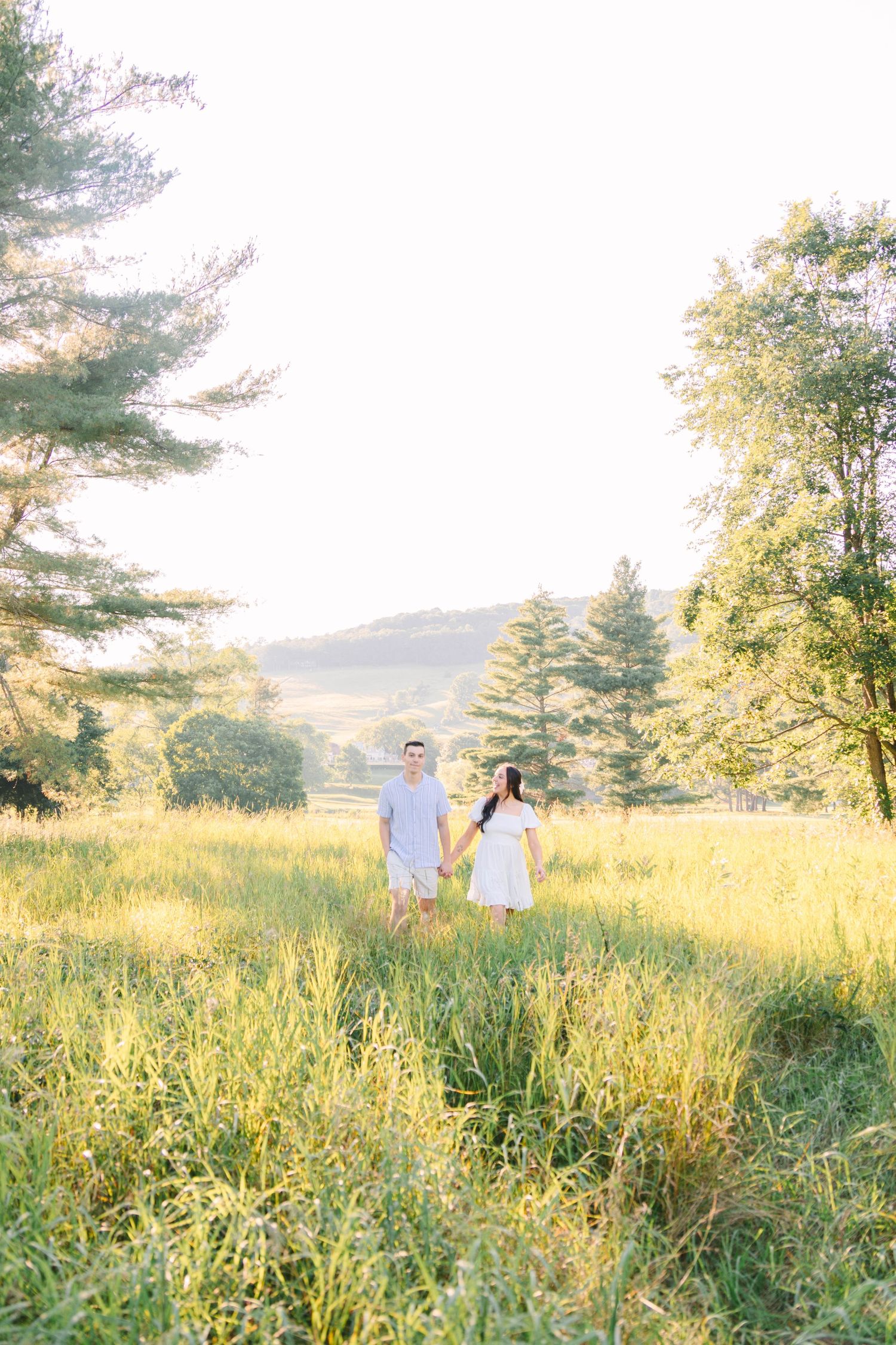 A couple holds hands in a sunlit green meadow surrounded by trees during a romantic outdoor engagement session.
