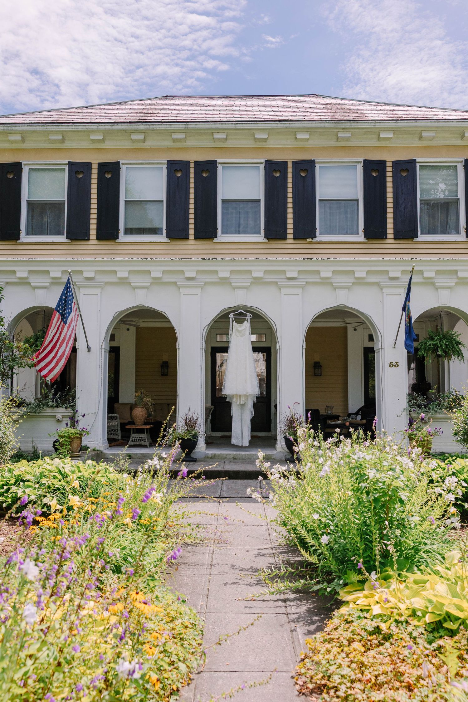 A white colonial mansion with black shutters, arched porch, American flag, and a wedding dress hanging, surrounded by colorful gardens.