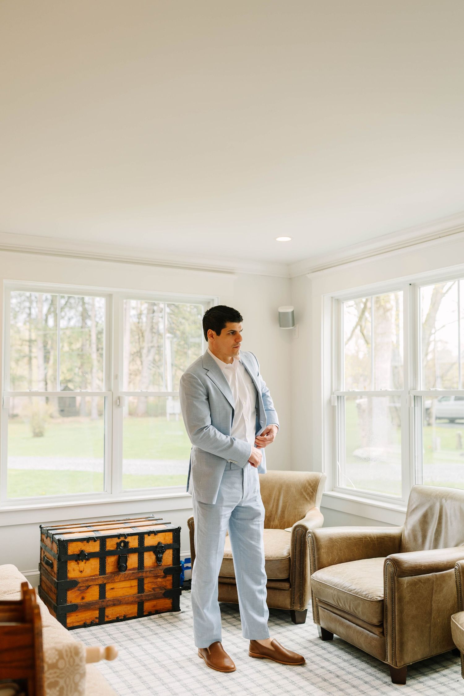 Groom buttoning light blue suit jacket in elegant sunroom with large windows.
