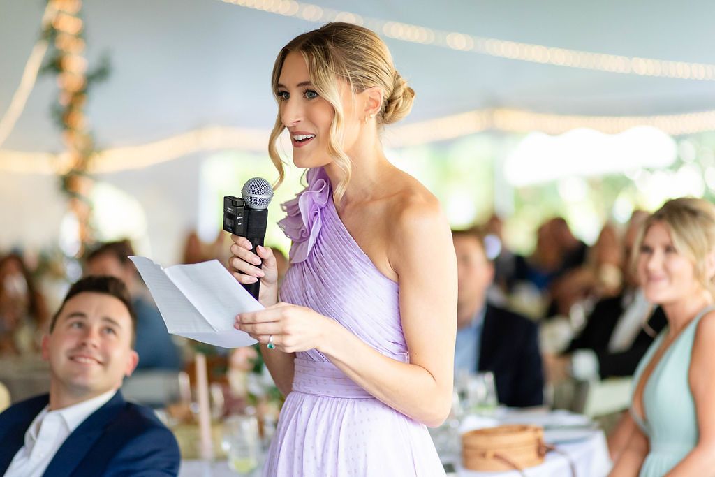 Bridesmaid in lavender dress delivers heartfelt speech with microphone at elegant tented wedding reception.