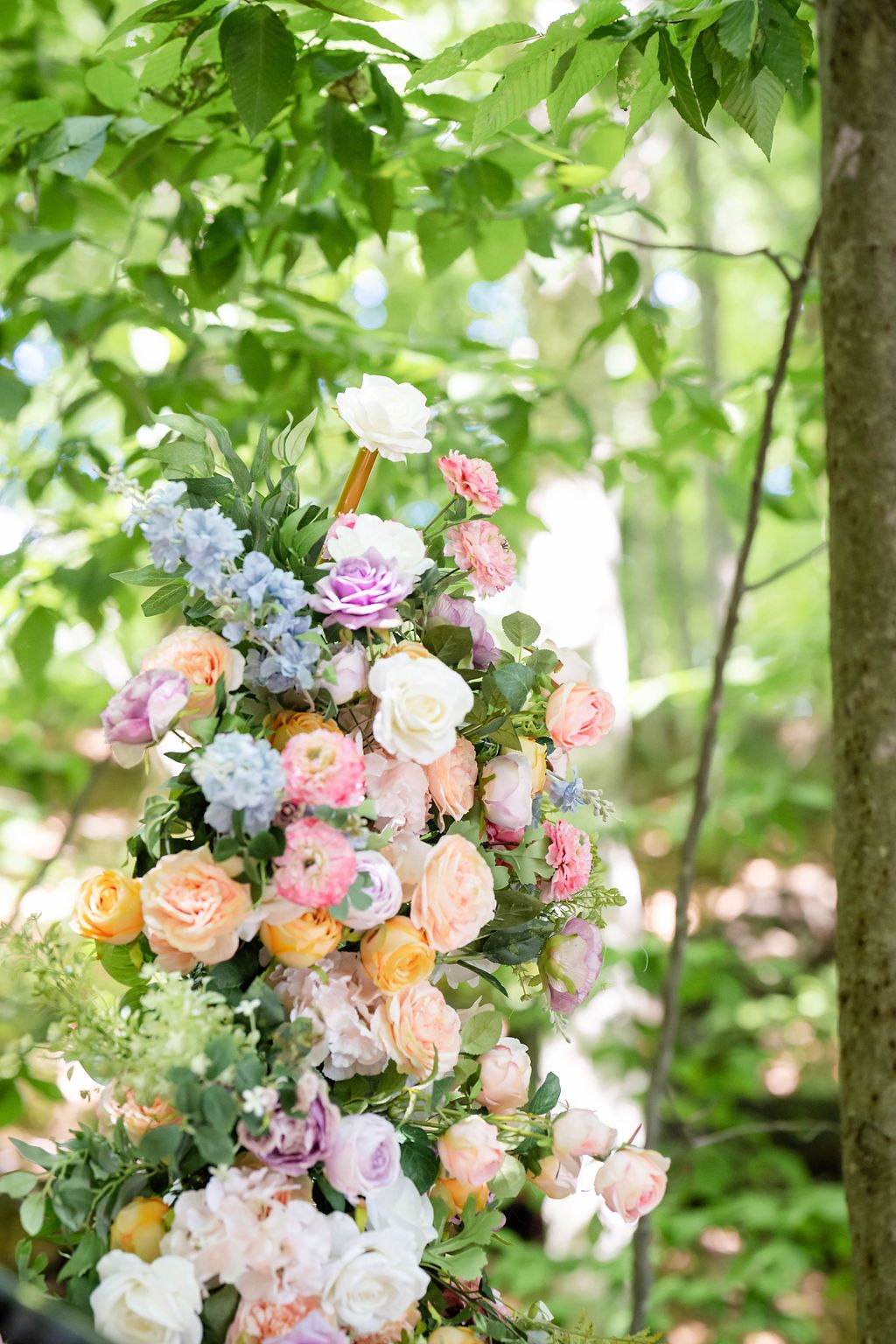 Lush floral arrangement of pastel roses and blooms decorating a wedding ceremony arch among green trees.