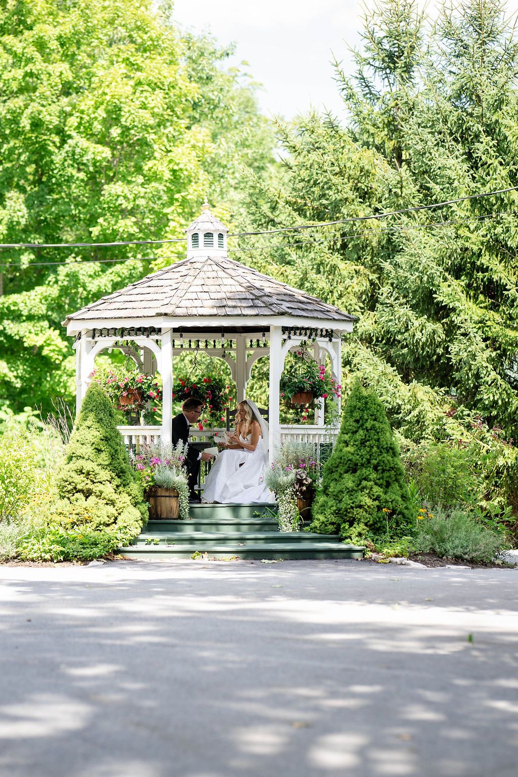 Bride and groom share a romantic moment in a white garden gazebo adorned with hanging flowers, surrounded by lush green trees.
