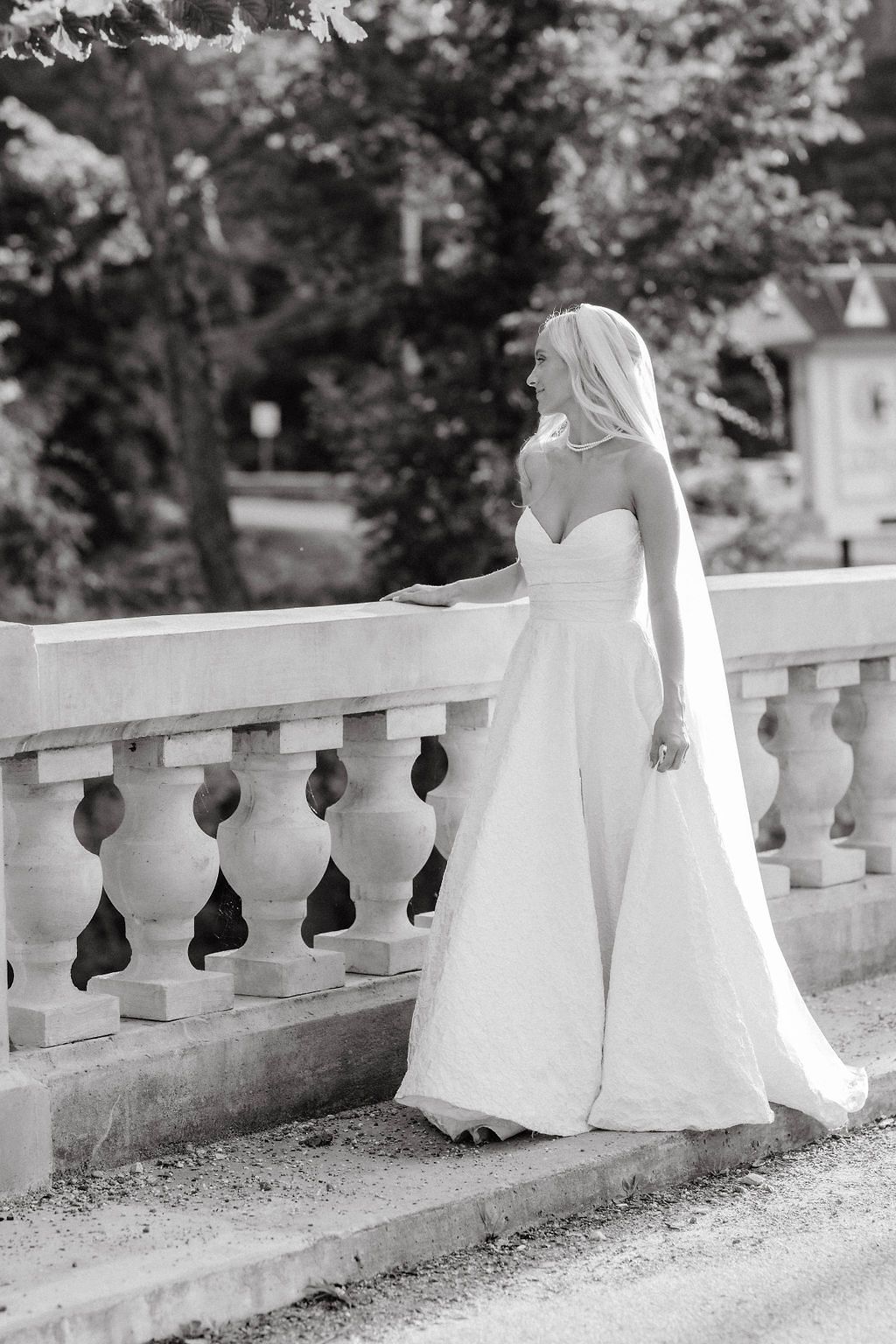 Black and white portrait of a bride in a strapless gown and veil posing gracefully beside a stone balustrade outdoors.