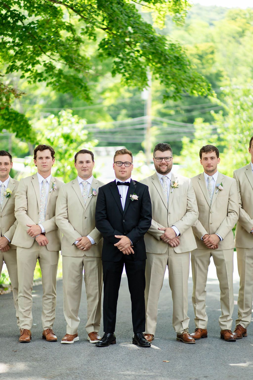 Groom in black tuxedo stands with groomsmen in tan suits outdoors under green trees.