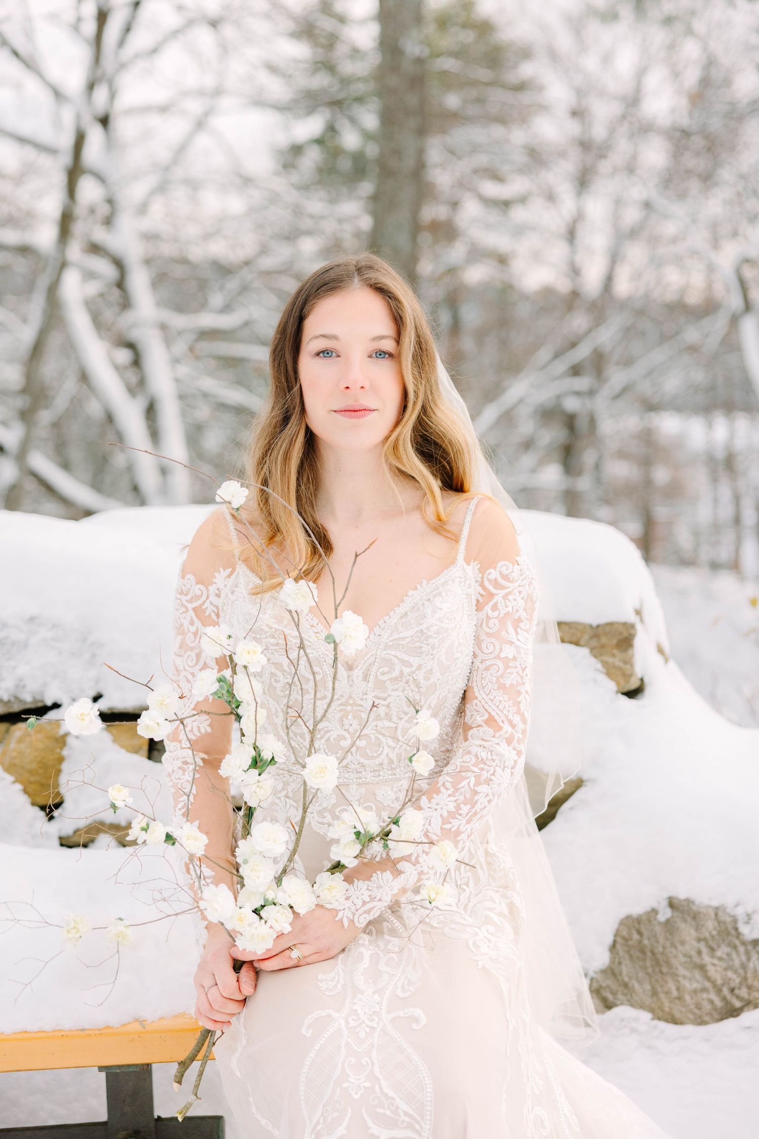 Bride in lace wedding dress sits on snow-covered bench holding white floral branch bouquet.