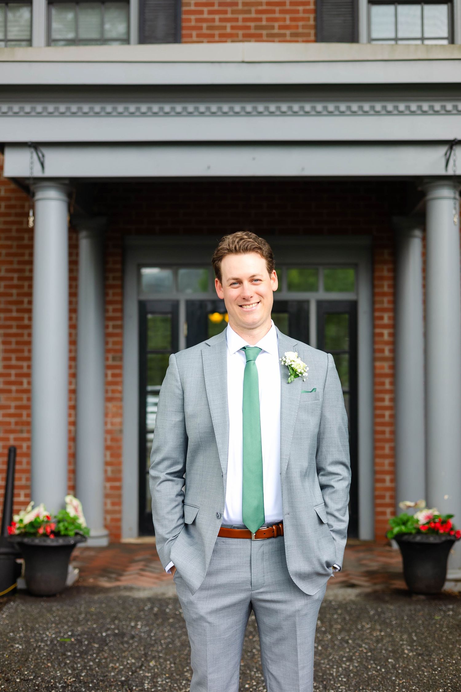 Smiling groom in gray suit with green tie and boutonniere stands outside brick wedding venue.