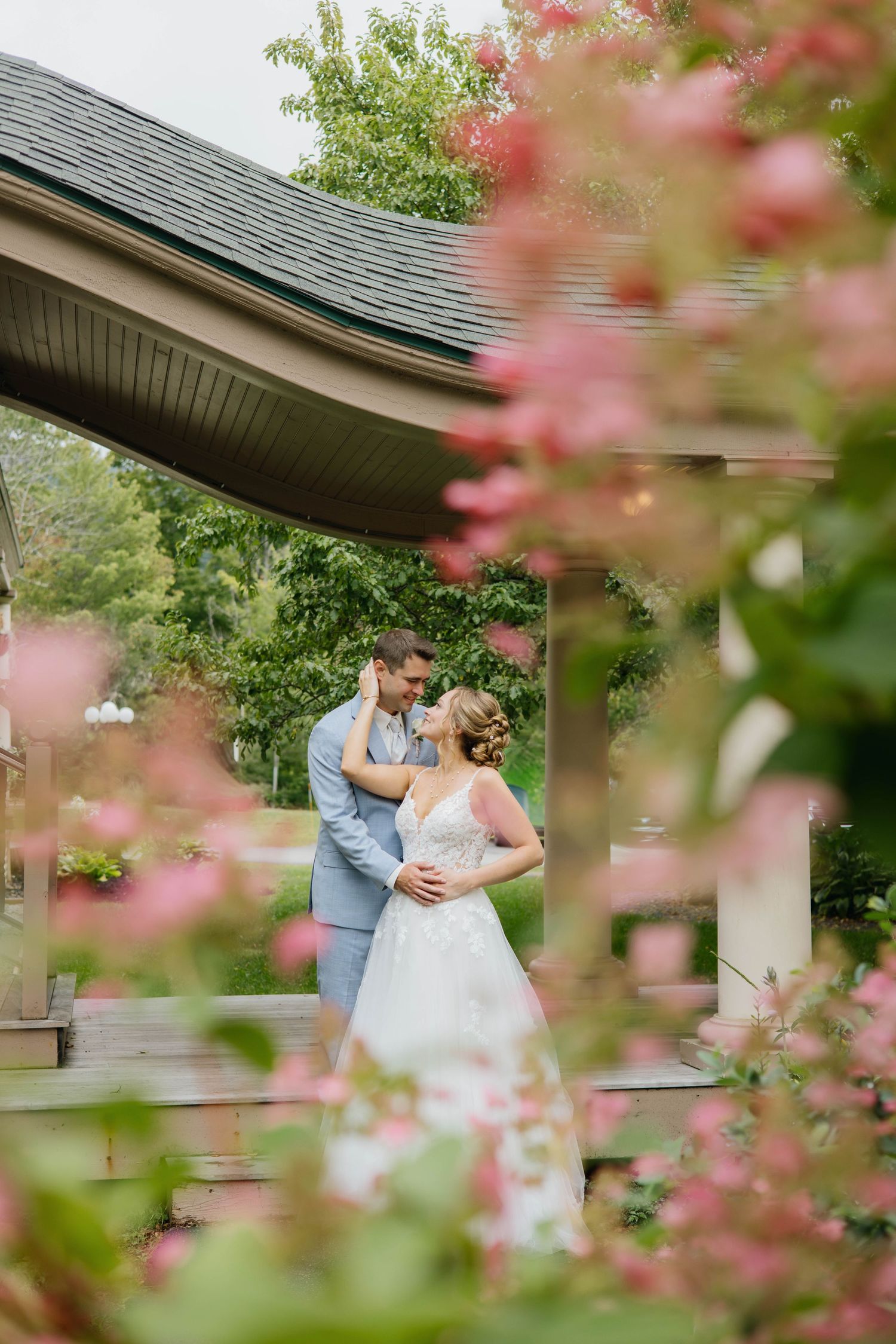 Bride and groom share a romantic moment under a gazebo, framed by blooming pink flowers in a lush garden setting.