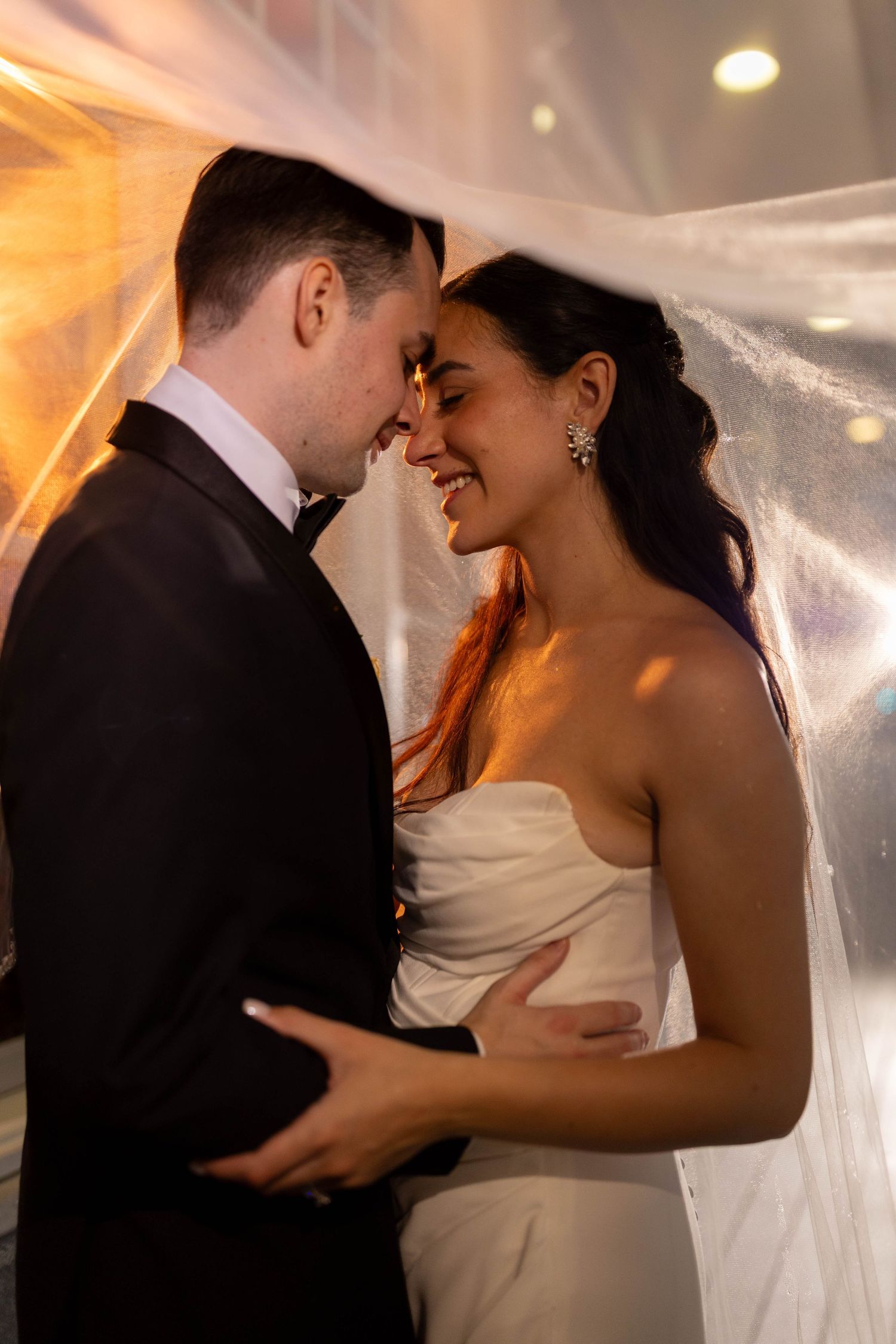 Bride and groom share an intimate moment under a veil with warm golden backlight at wedding reception.