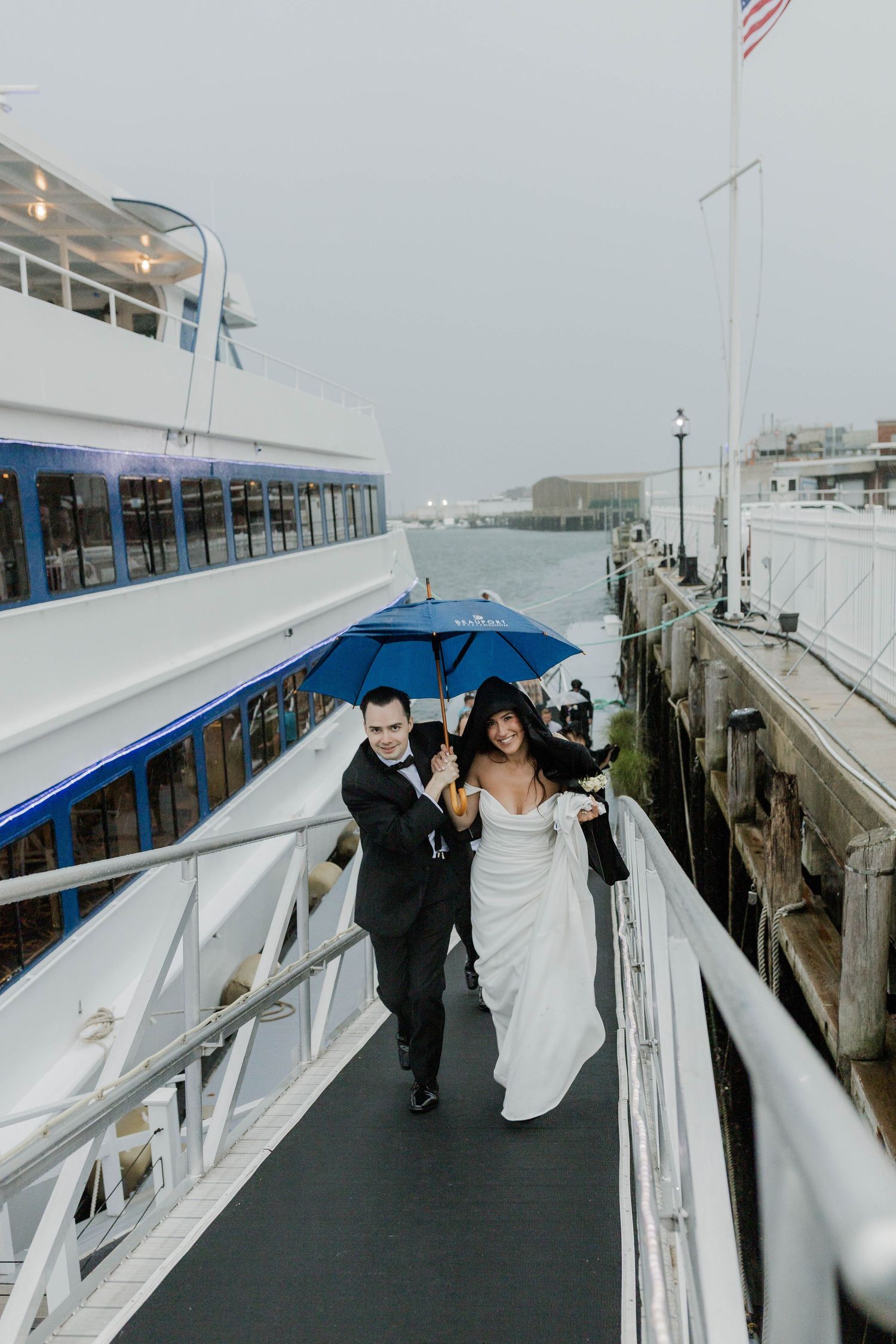 Bride and groom sharing a blue umbrella walking along a rainy waterfront dock near a yacht.