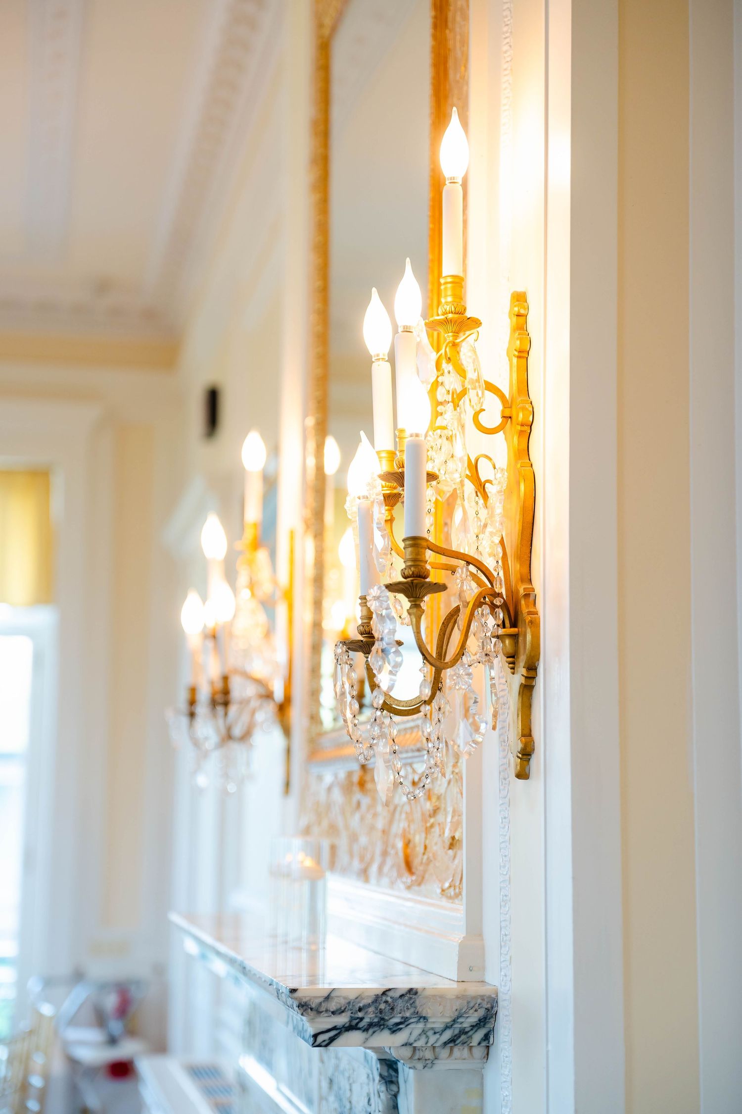 Row of ornate gold crystal candelabra wall sconces glowing in elegant white wedding venue hallway.