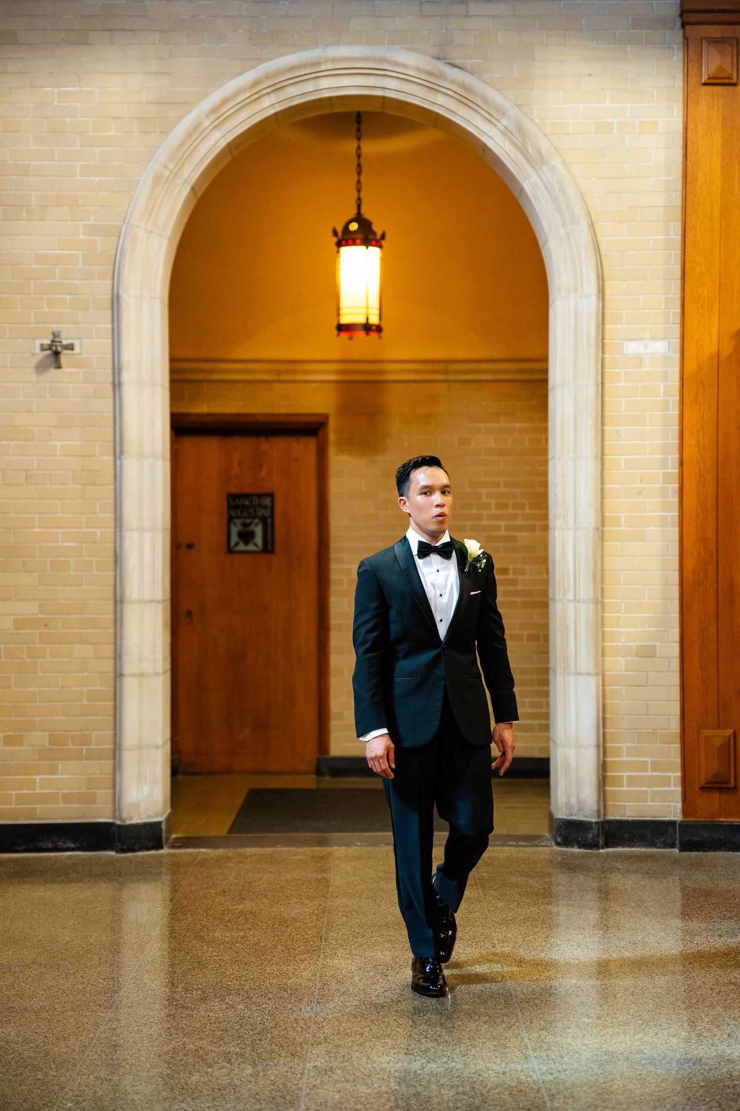 Groom in black tuxedo with boutonniere walking through a stone archway in a classic wedding venue.