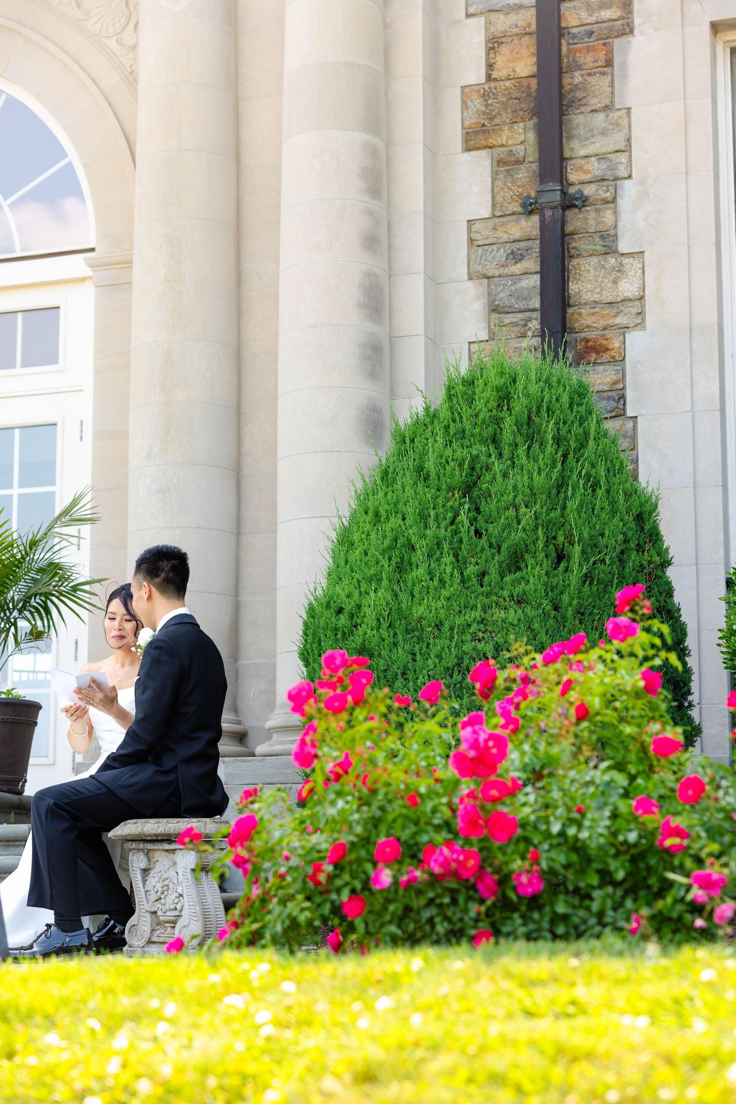 Bride and groom sit together among vibrant pink roses outside elegant stone venue.