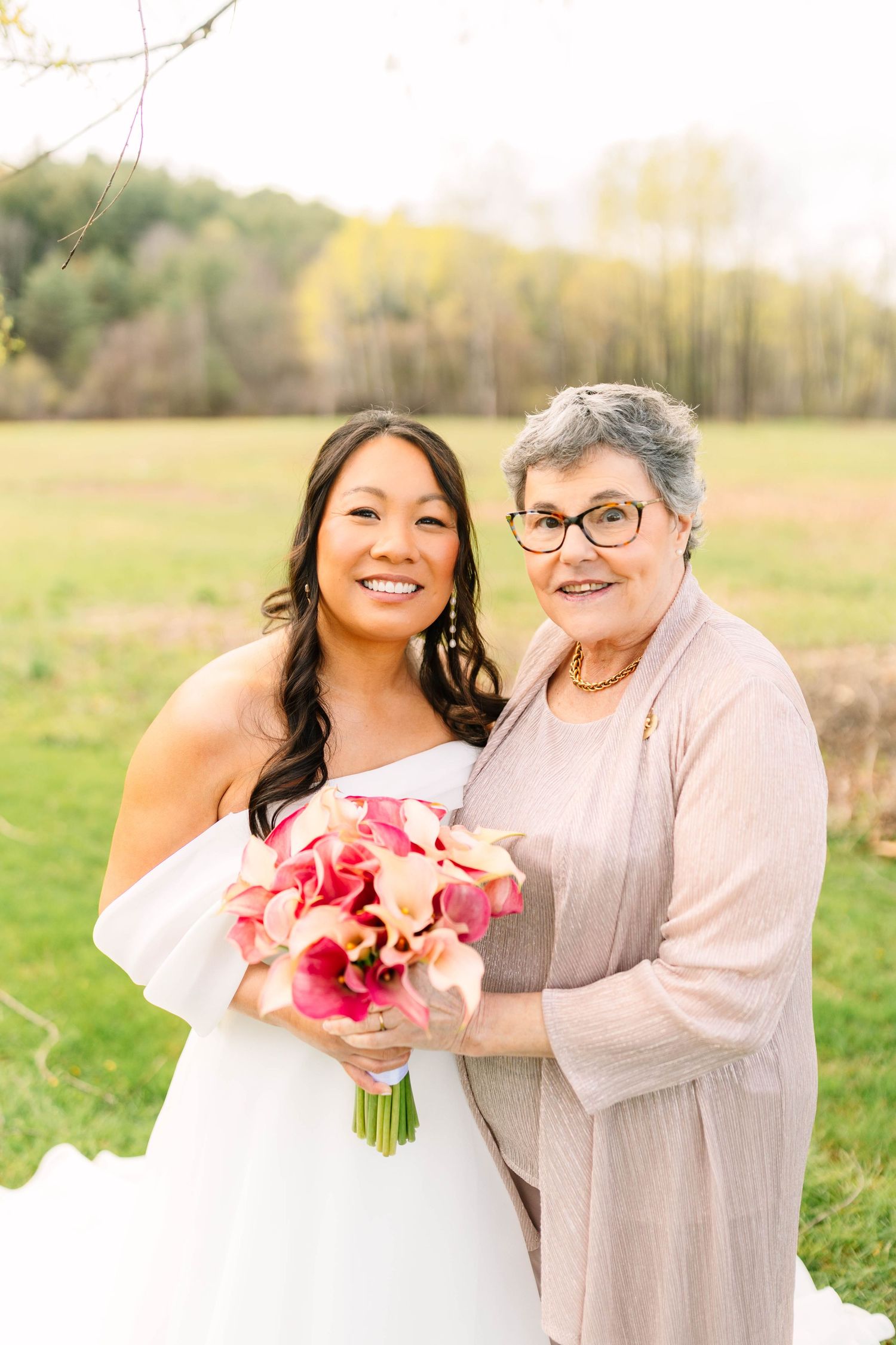 Bride in white gown holds pink calla lily bouquet with older woman in taupe cardigan at outdoor wedding.