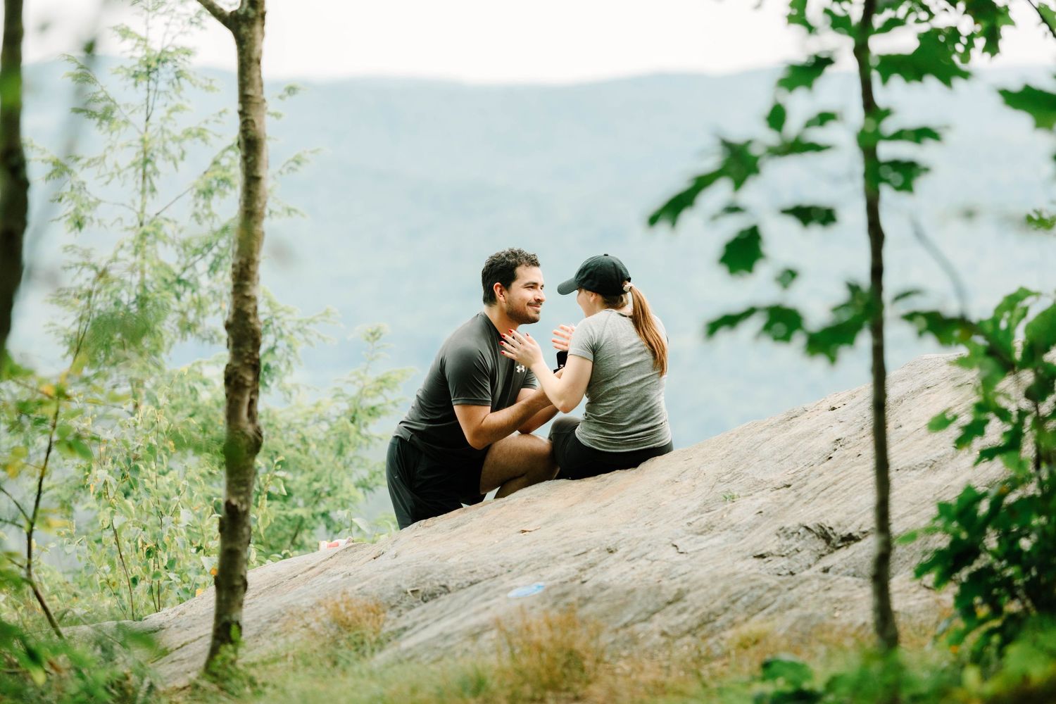 Couple shares intimate engagement moment seated on large rock surrounded by lush green forest trees.