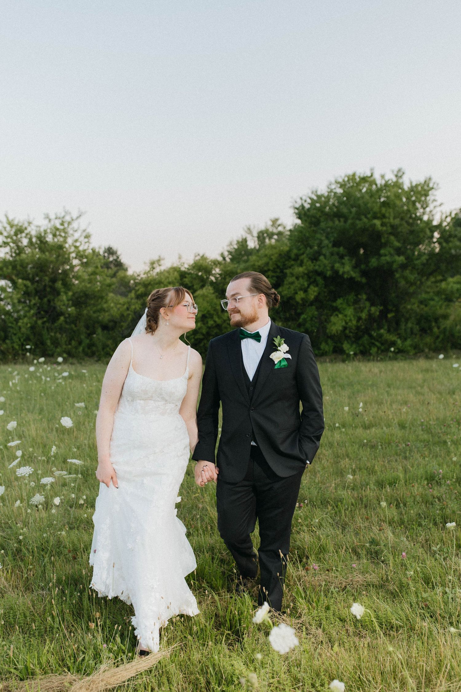 Bride and groom walk hand-in-hand through a green meadow, exchanging smiles in golden hour light.