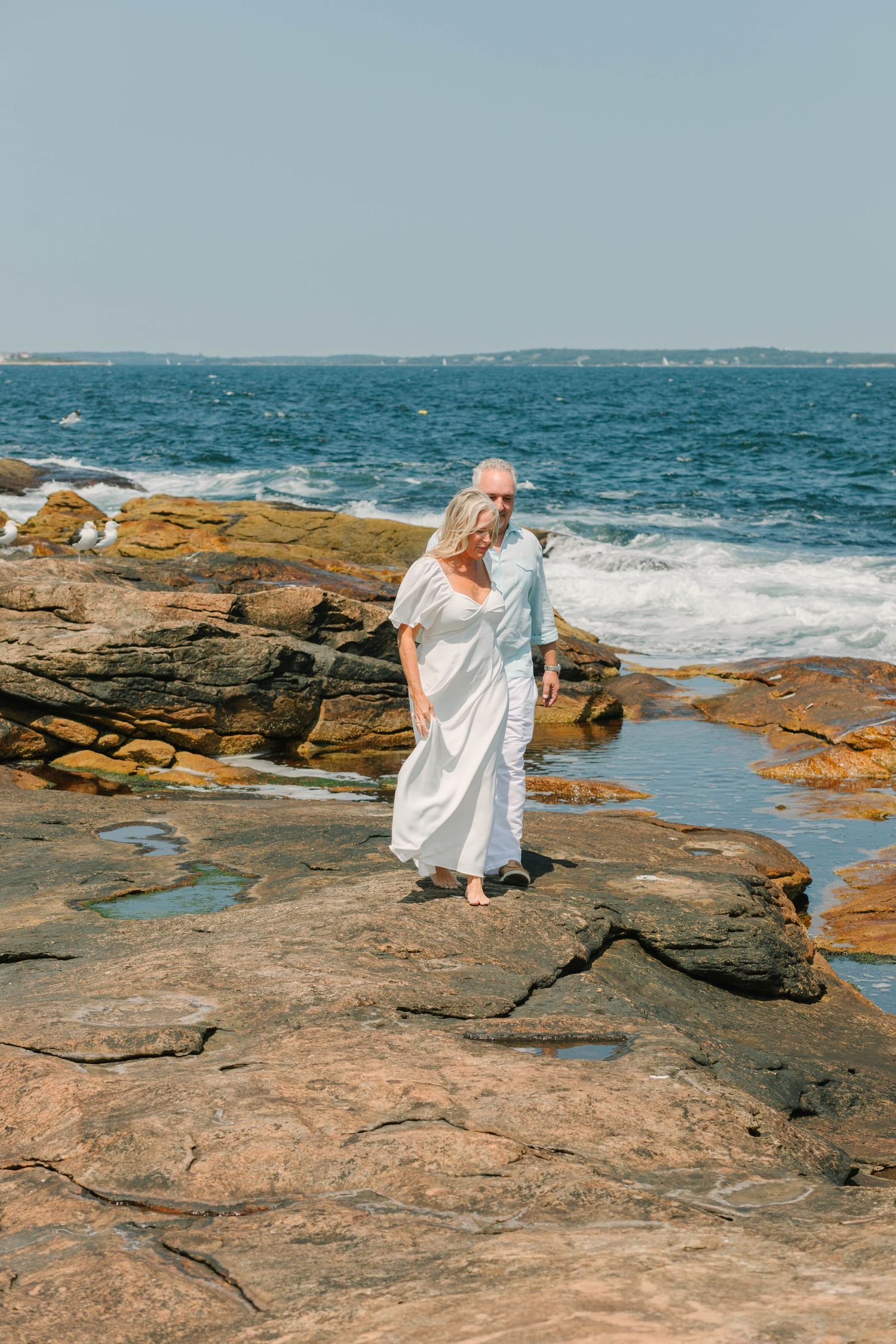 Mature couple walking hand in hand on rocky shoreline with ocean waves crashing behind them on clear day.