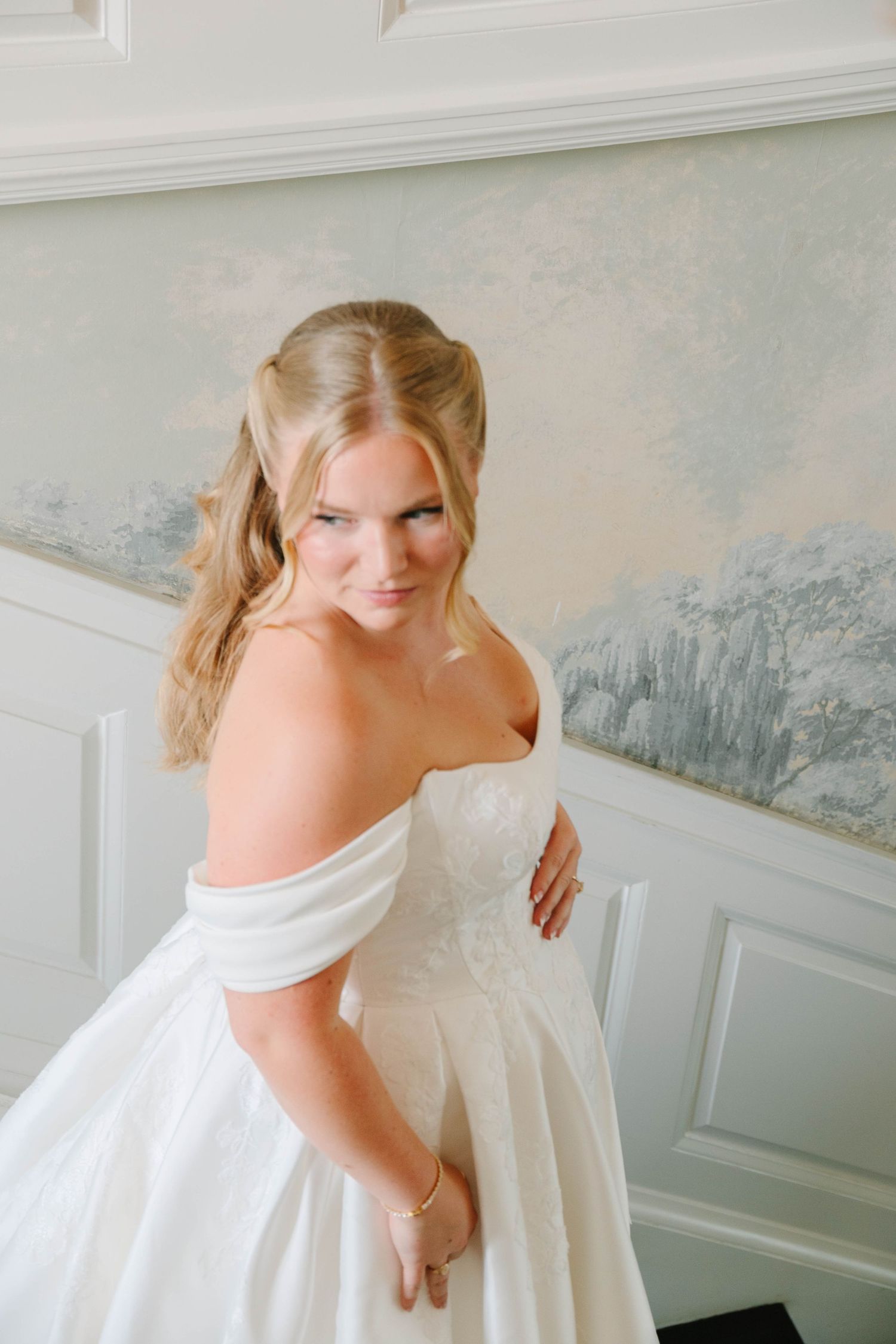Blonde bride in off-shoulder white ball gown poses elegantly in a softly lit room with scenic wallpaper.