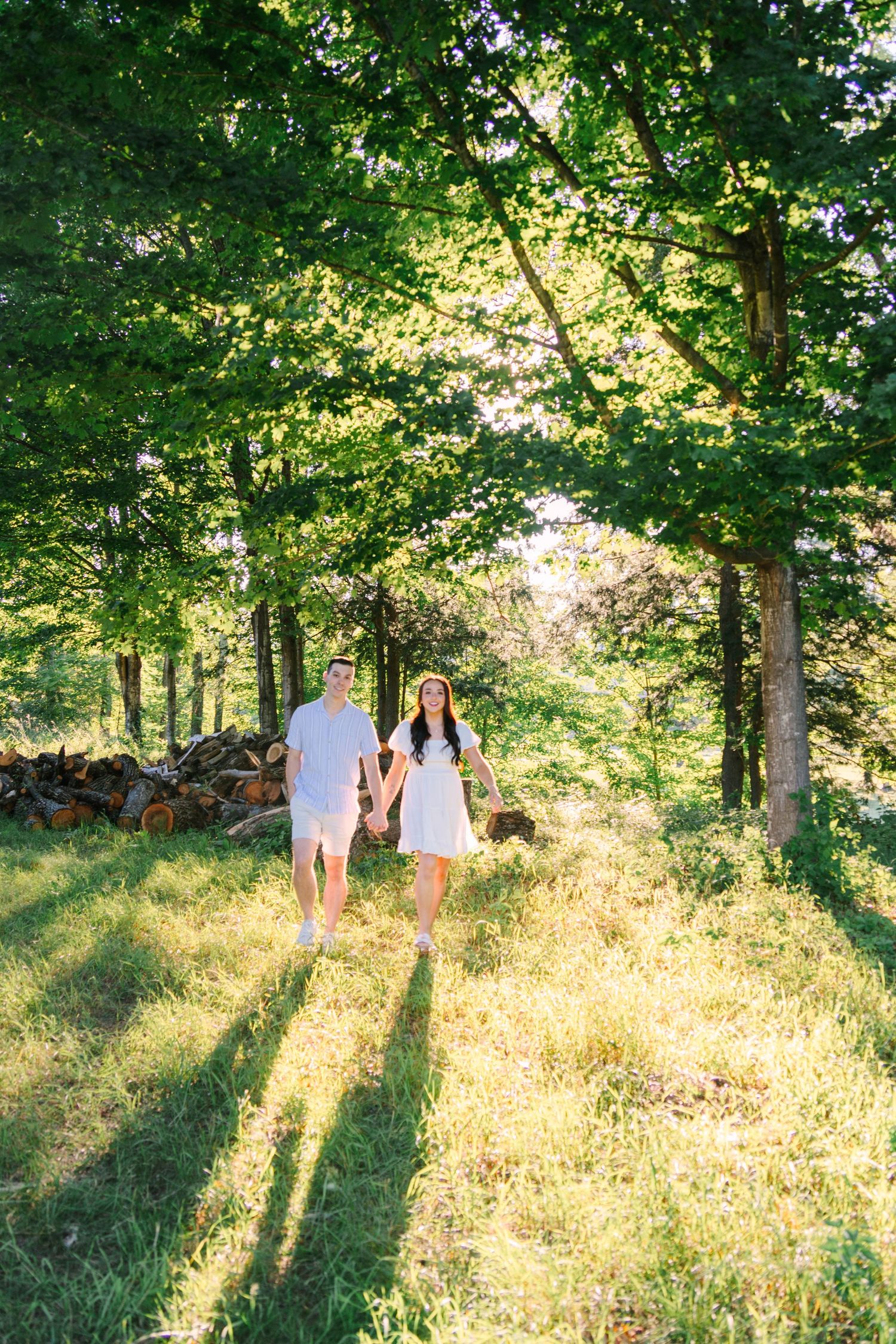 Couple holding hands walking through sun-dappled green tree-lined path outdoors.