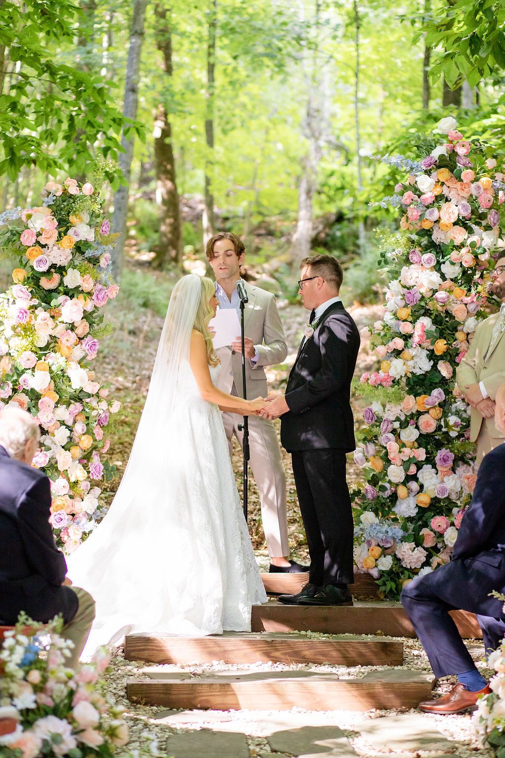 Bride and groom exchanging vows at stunning outdoor forest wedding ceremony with pastel floral columns.