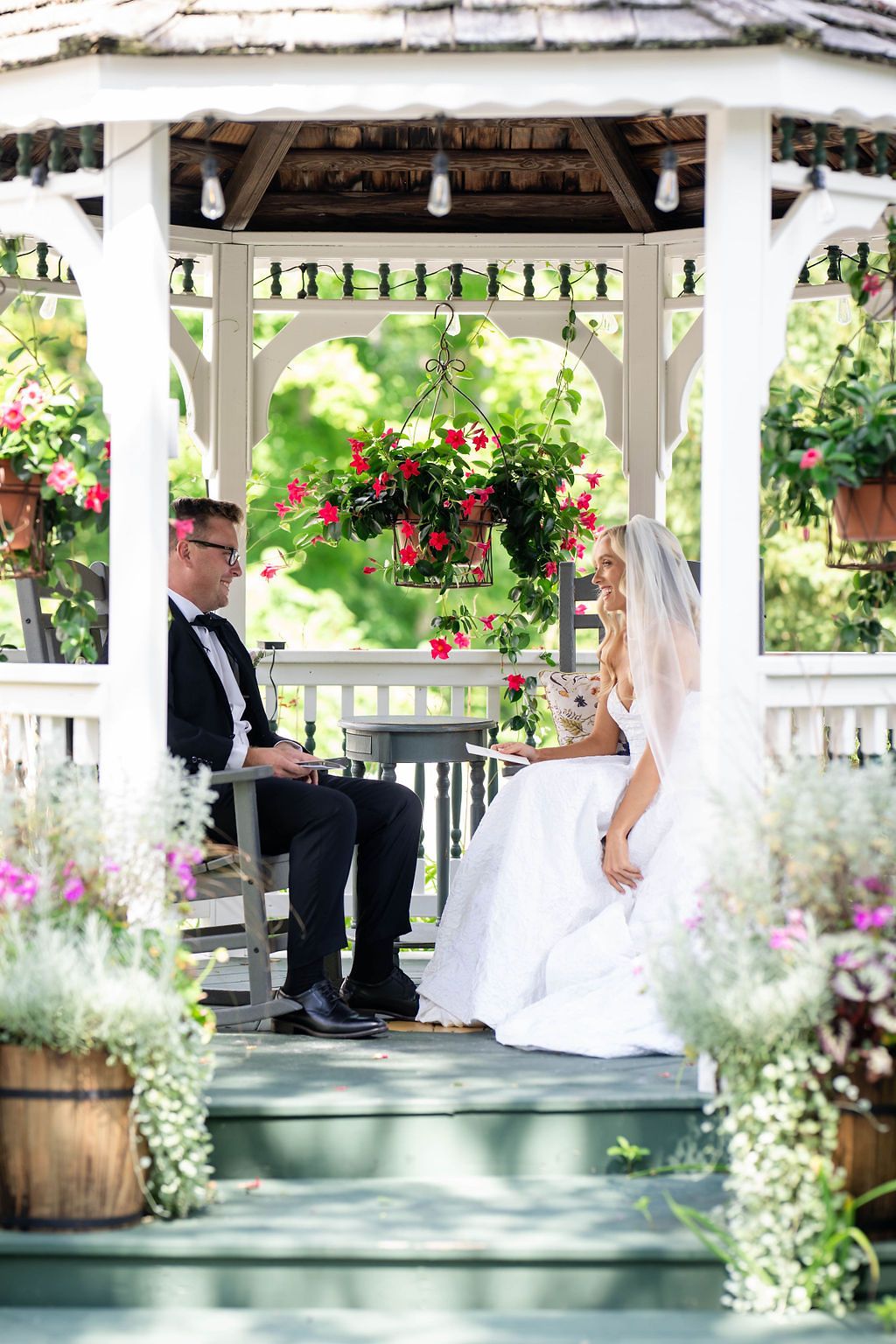 Bride and groom sitting together in a white gazebo decorated with hanging flower baskets and surrounded by lush greenery.