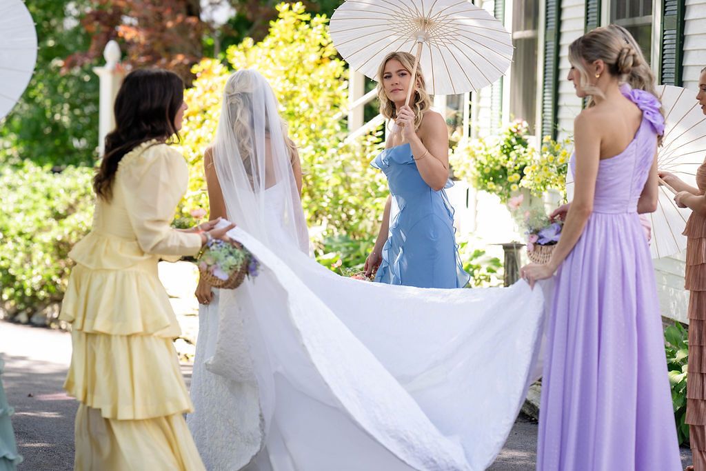 Bridesmaids in colorful dresses help bride with long veil and parasol in sunny outdoor garden setting.