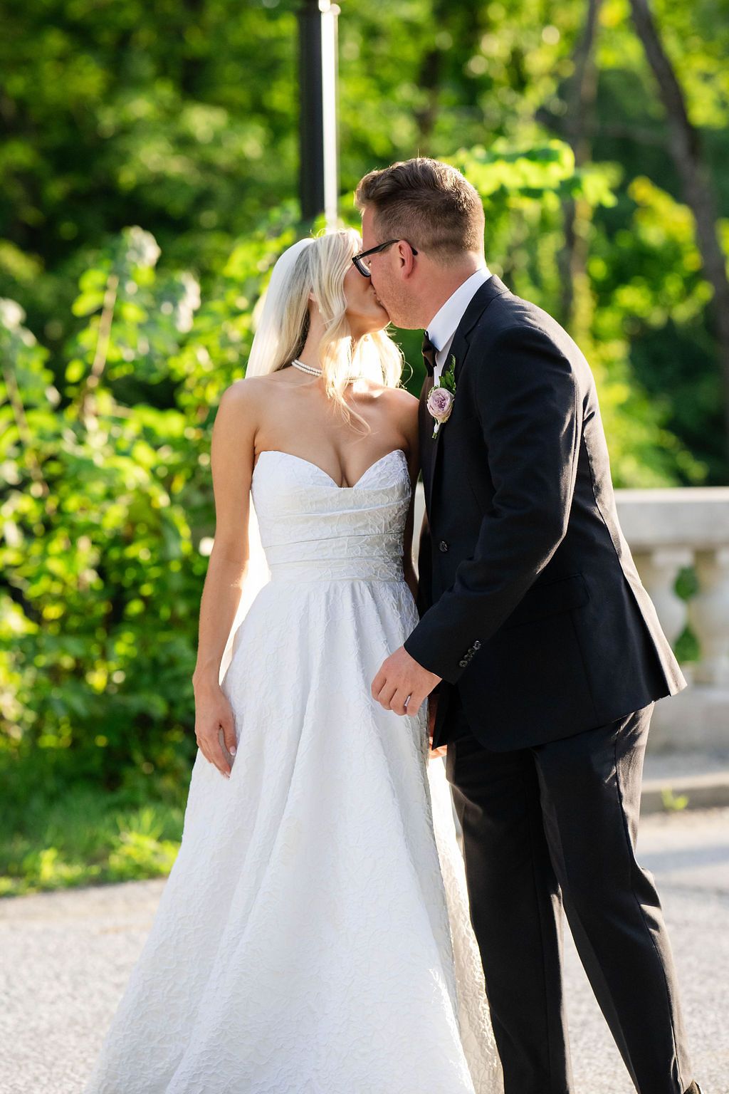 Bride and groom kissing outdoors on a sunny day, bride in white strapless gown with veil, groom in black suit.
