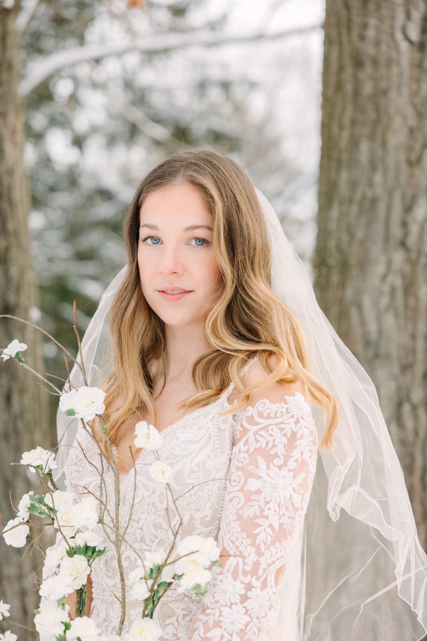 Close portrait of bride in lace gown and veil holding white floral branches in winter forest.