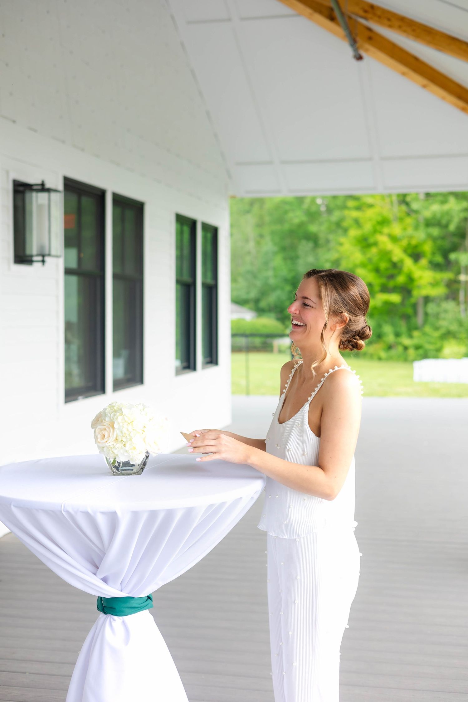 Smiling bride in white pajamas arranging floral centerpiece on cocktail table with green ribbon outdoors.