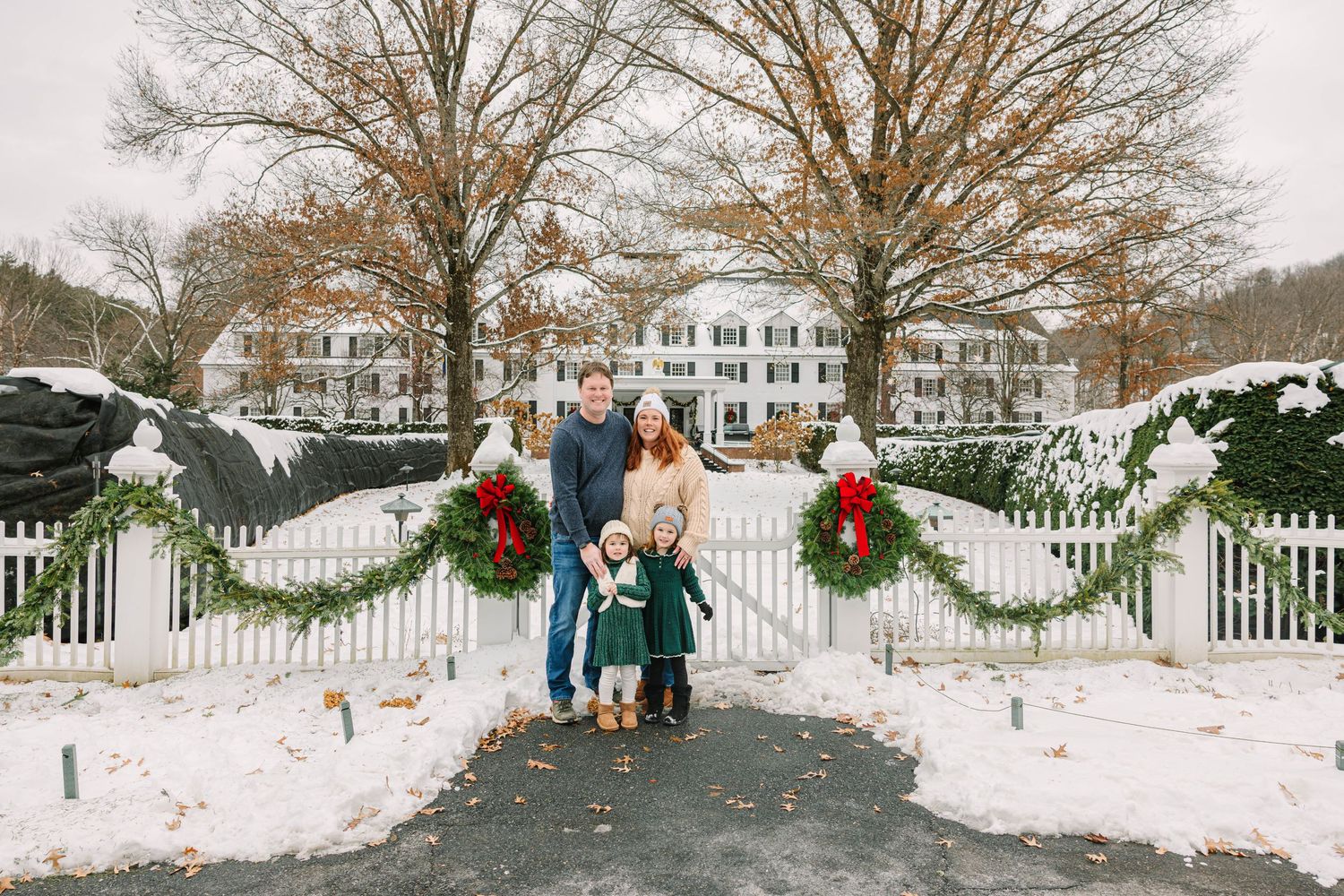 A family of four poses at a snow-covered white fence entrance decorated with Christmas wreaths and red holiday bows.