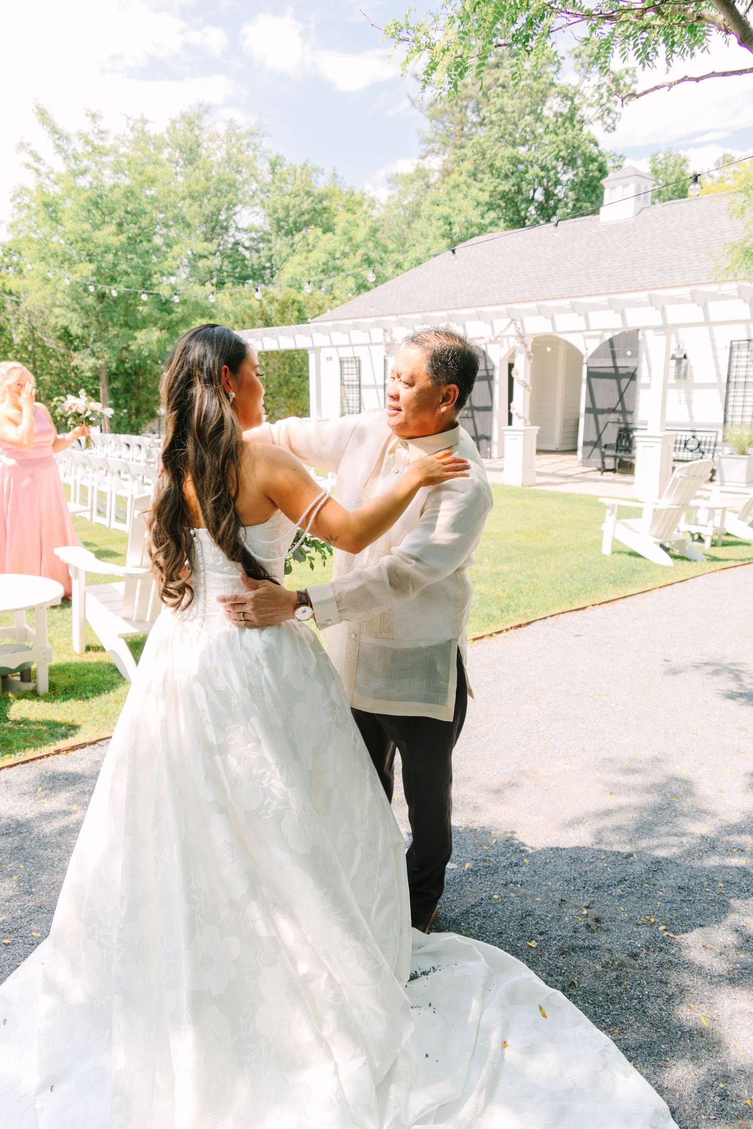 Bride and father share a tender first look moment, embracing outdoors at a sunny wedding venue with white chairs.