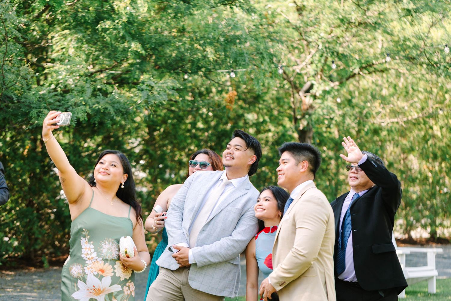 Wedding guests taking group selfie outdoors among lush green trees.