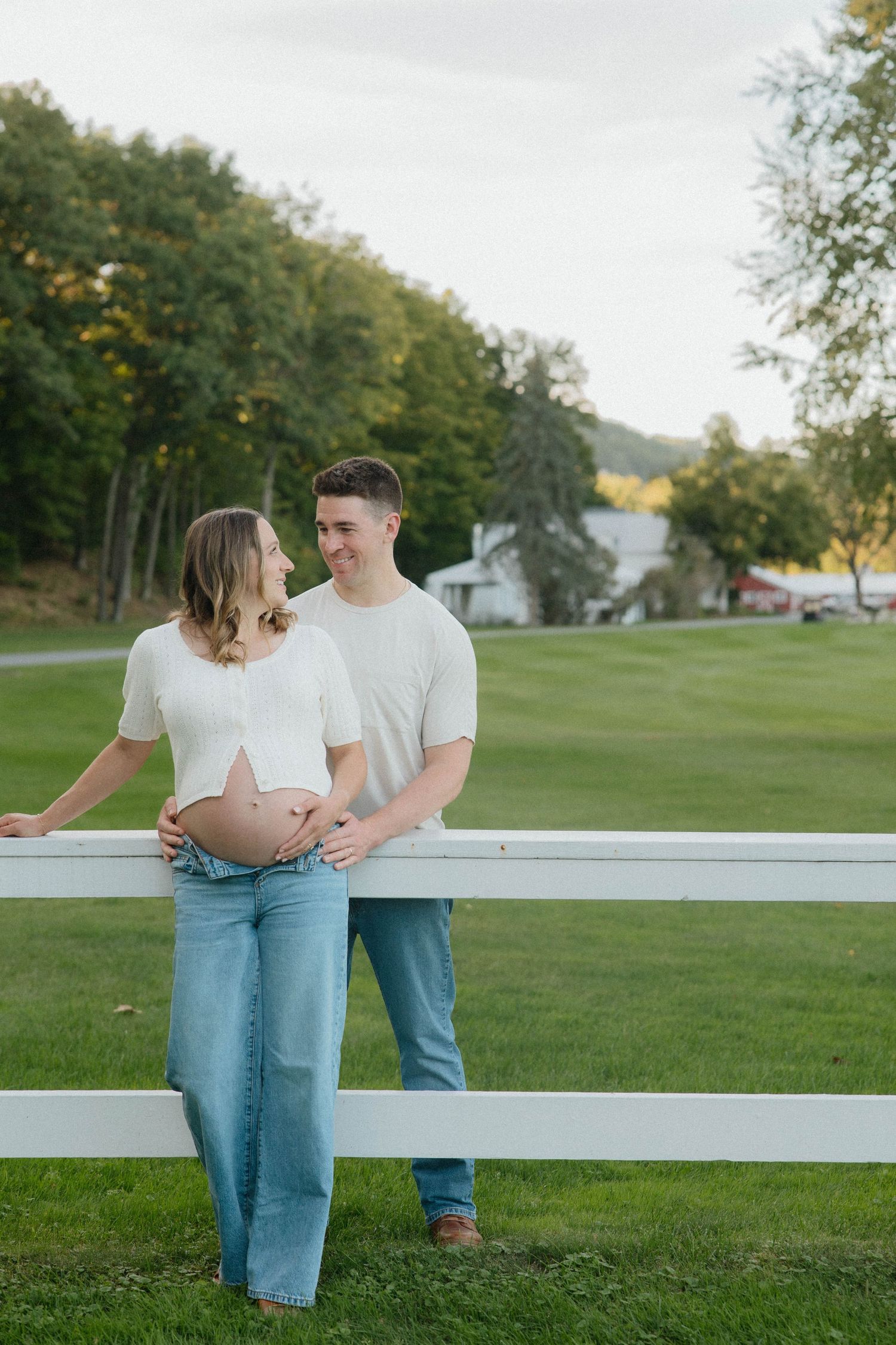 Pregnant couple in white tops and jeans leaning against a white fence outdoors, smiling at each other on a green lawn.