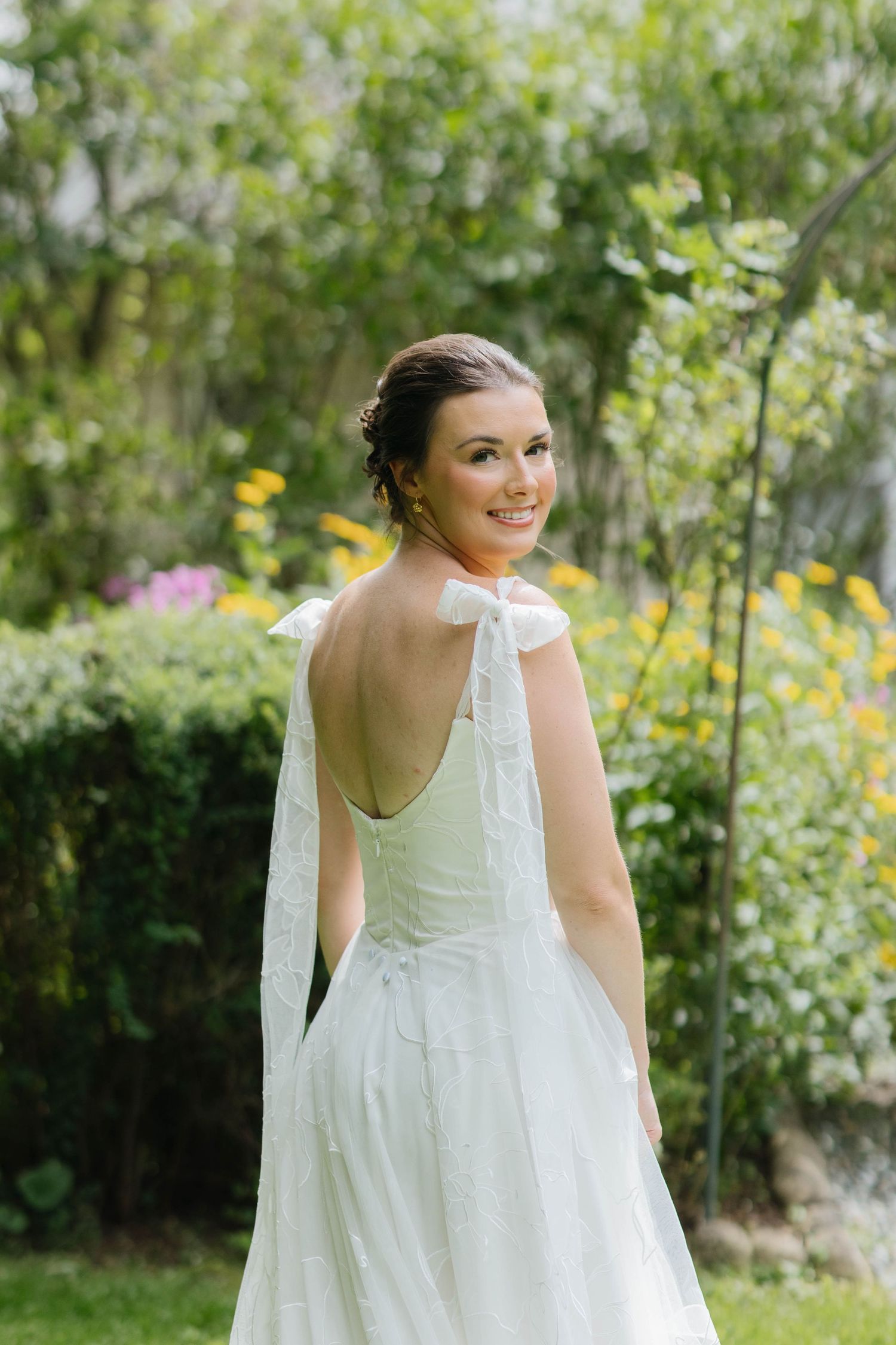 Smiling bride in a white backless wedding gown with bow detail, posing in a lush garden with colorful flowers.