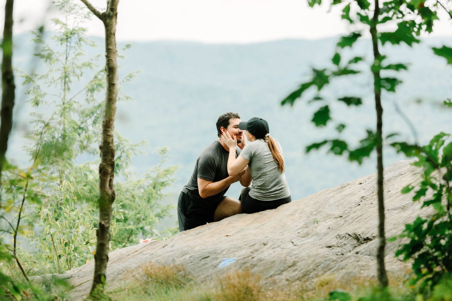 Couple kissing on a rocky overlook surrounded by lush green trees with misty mountain views behind them.