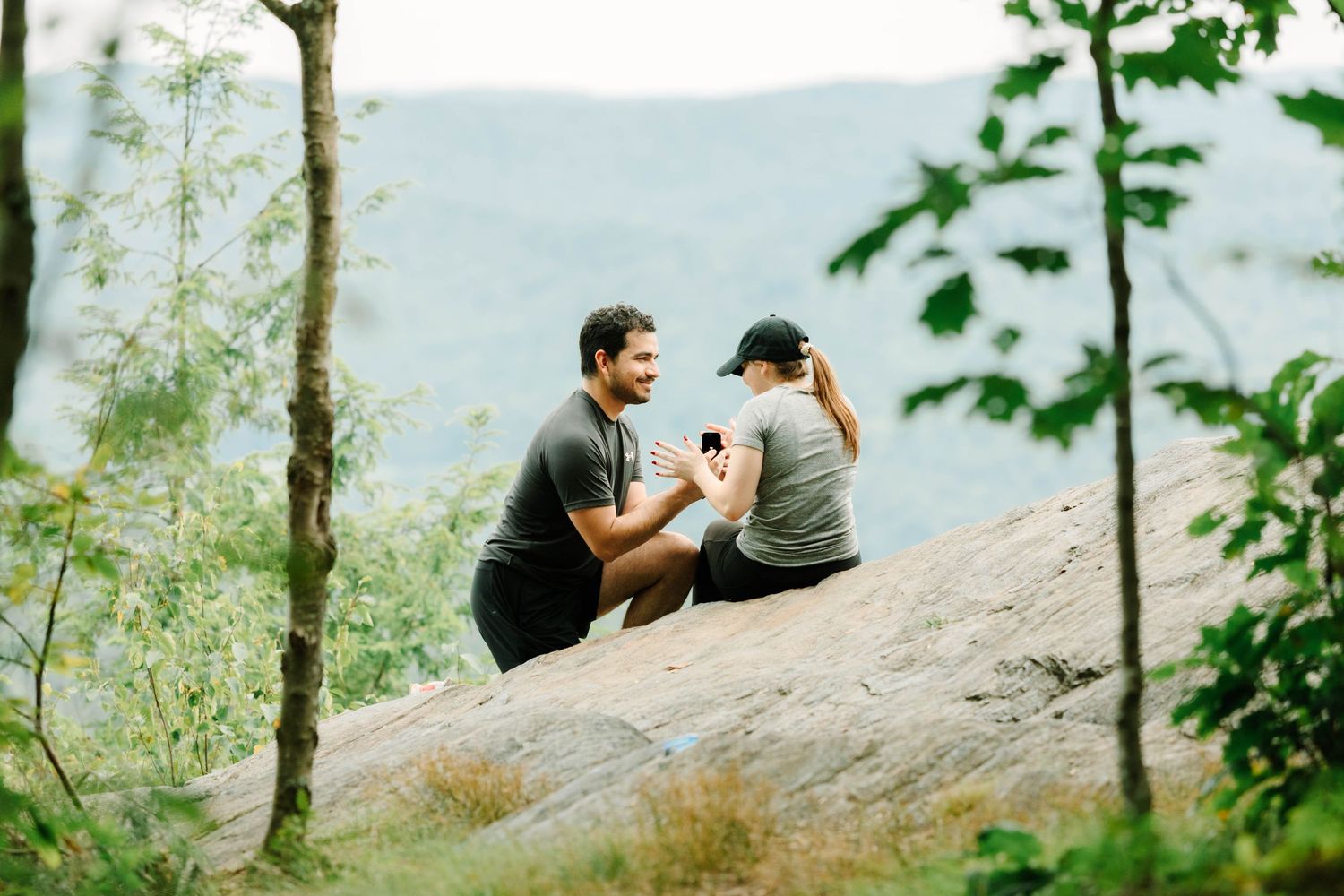 Man proposing to woman on a rocky overlook surrounded by lush green trees and mountains in the background.