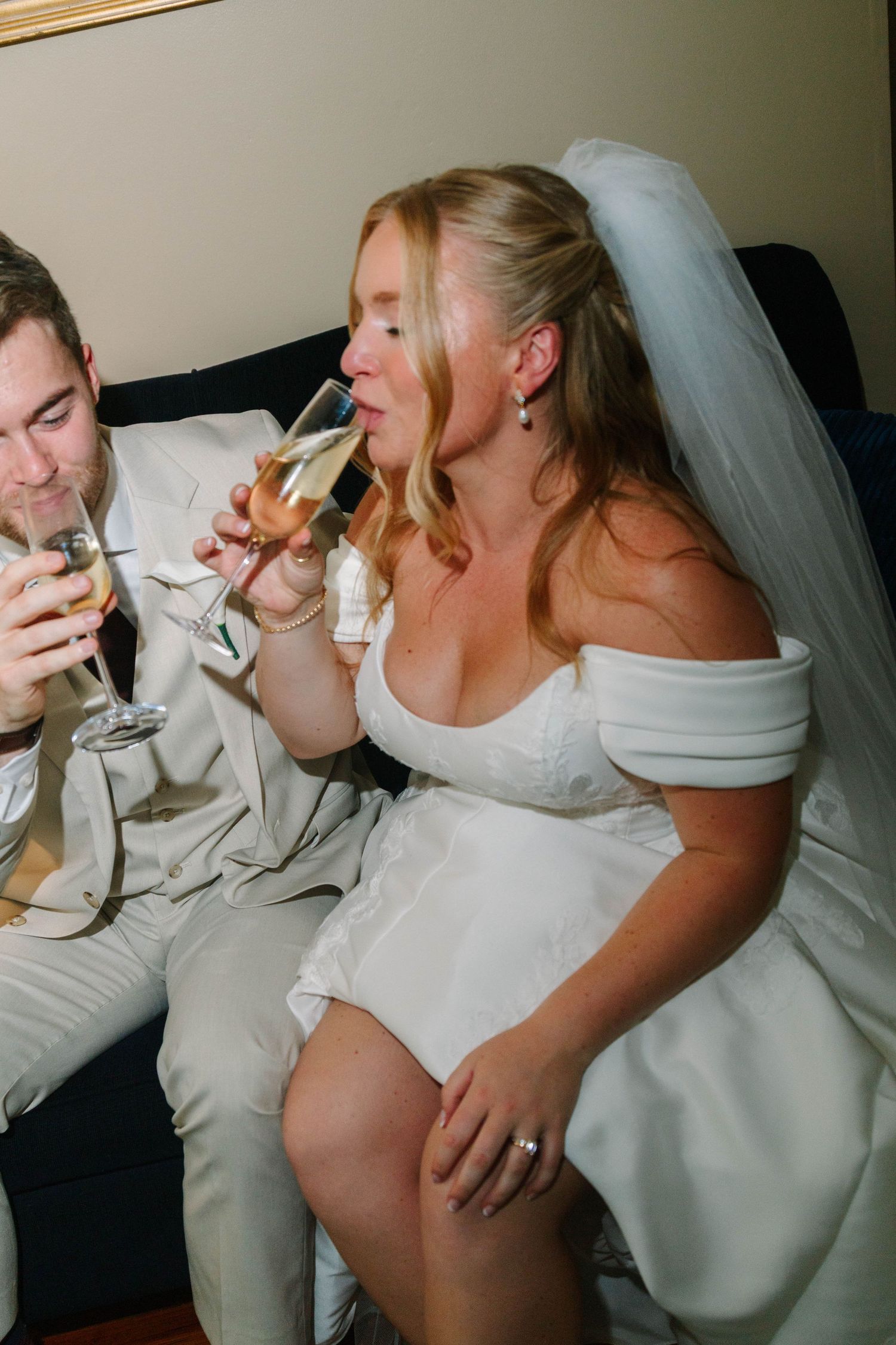 Bride and groom toasting with champagne glasses while seated on a sofa during their wedding reception.