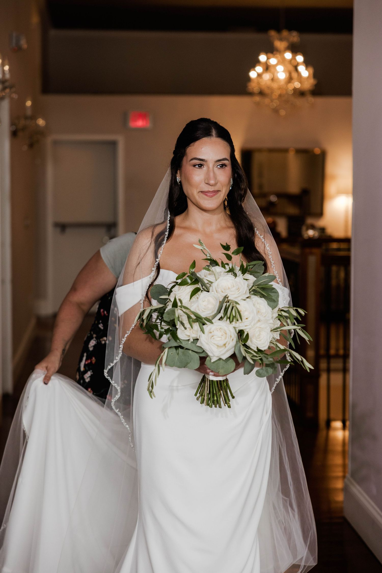 Beautiful bride in white off-shoulder gown holding white rose bouquet with veil indoors.